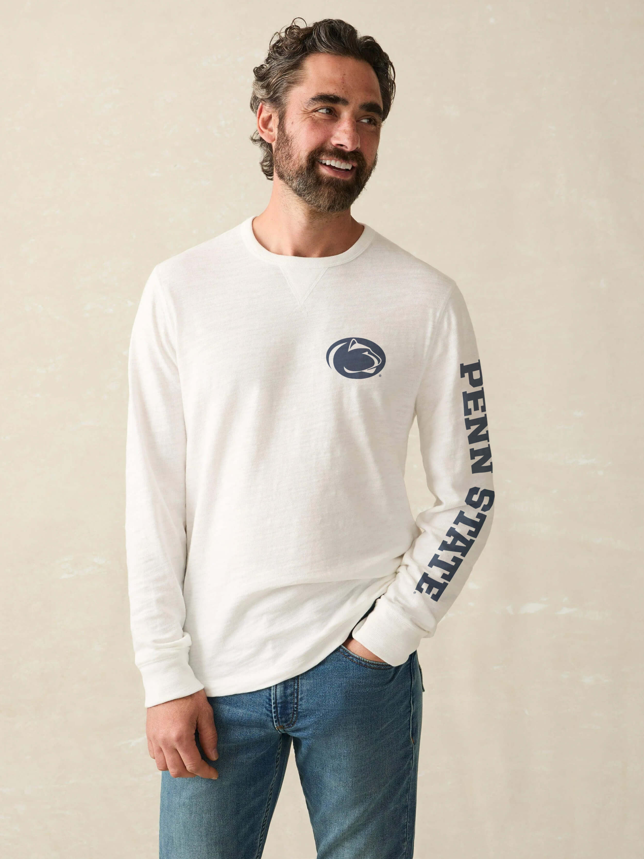 Man wearing white long sleeve t-shirt with Penn State logo on the chest and text down the left sleeve against a beige background