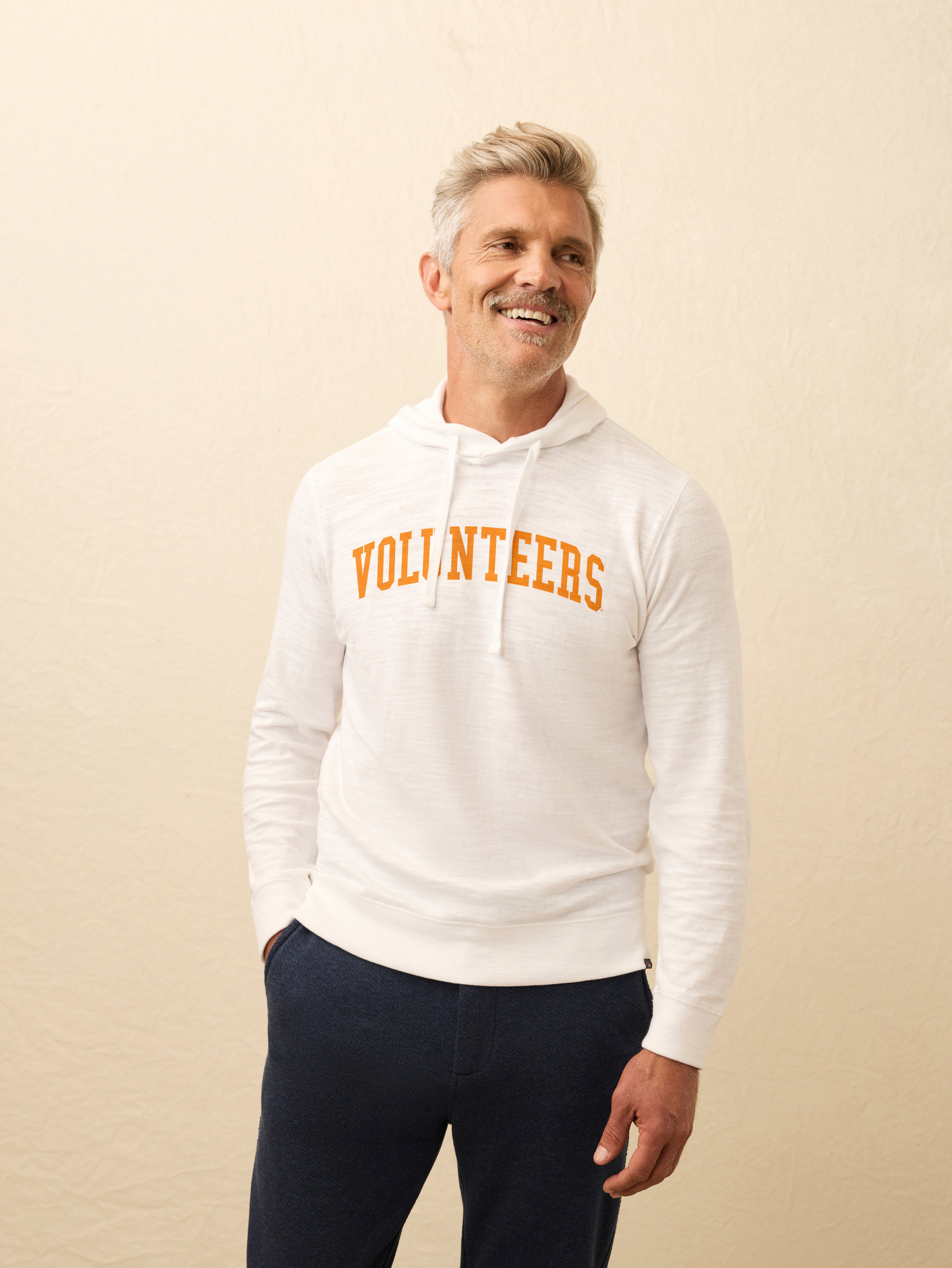 Man is wearing a white hoodie with University of Tennessee 'Volunteers' graphic on the center chest against a plain background