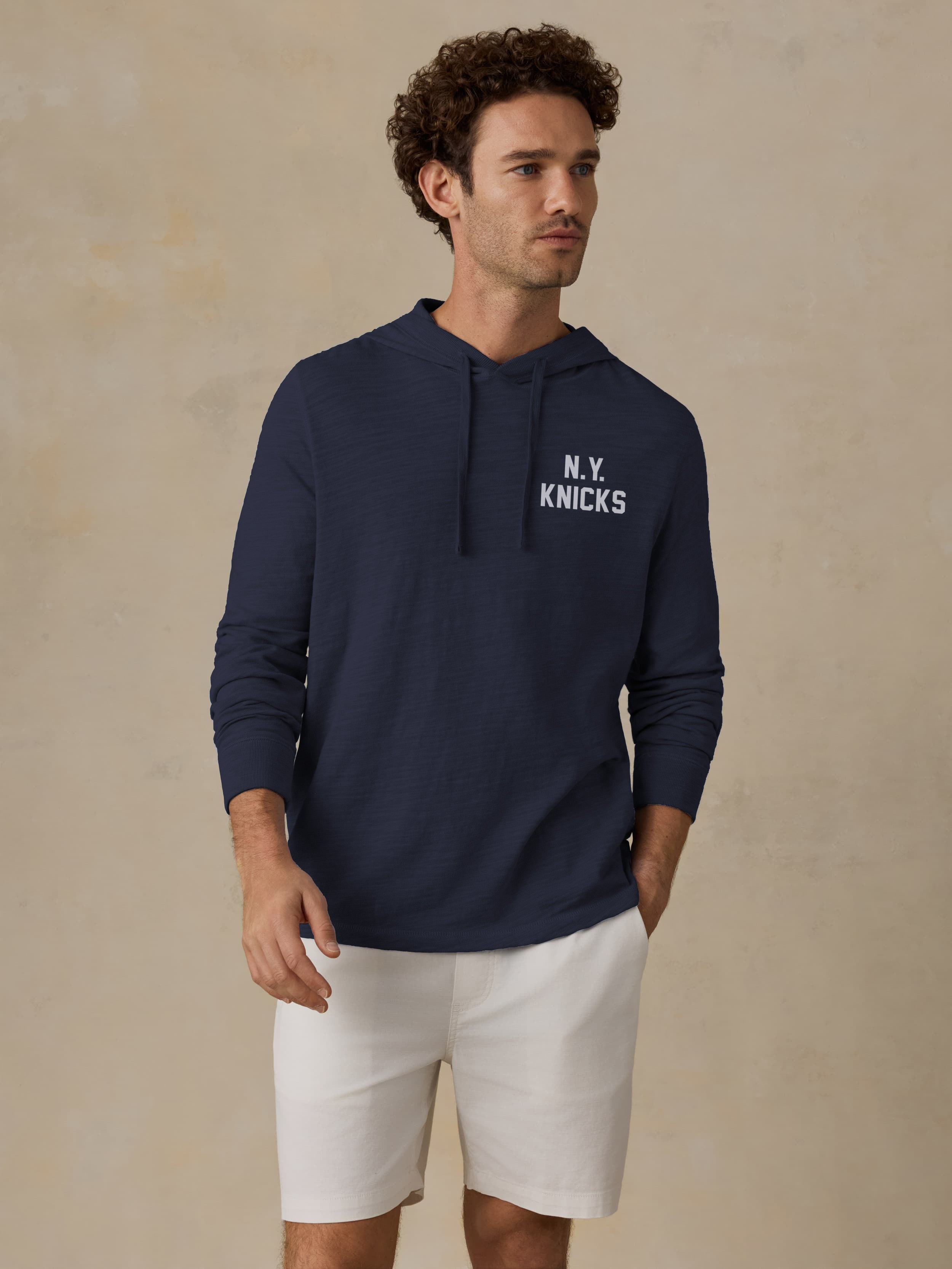 Man wearing a navy blue NY Knicks graphic text on the left chest against a beige background