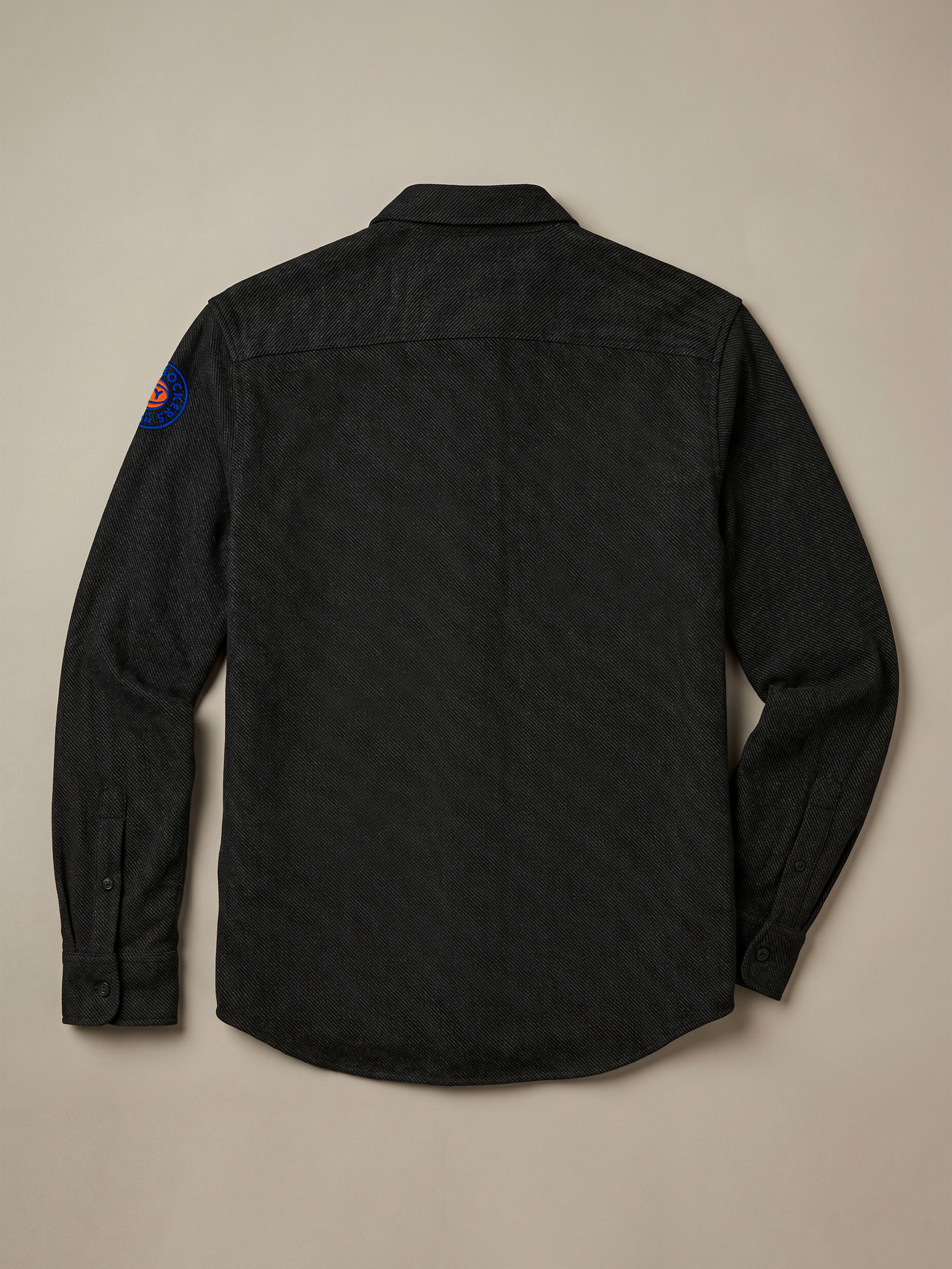 Black shirt with NY Knicks embroidered graphic on the left shoulder against a beige background
