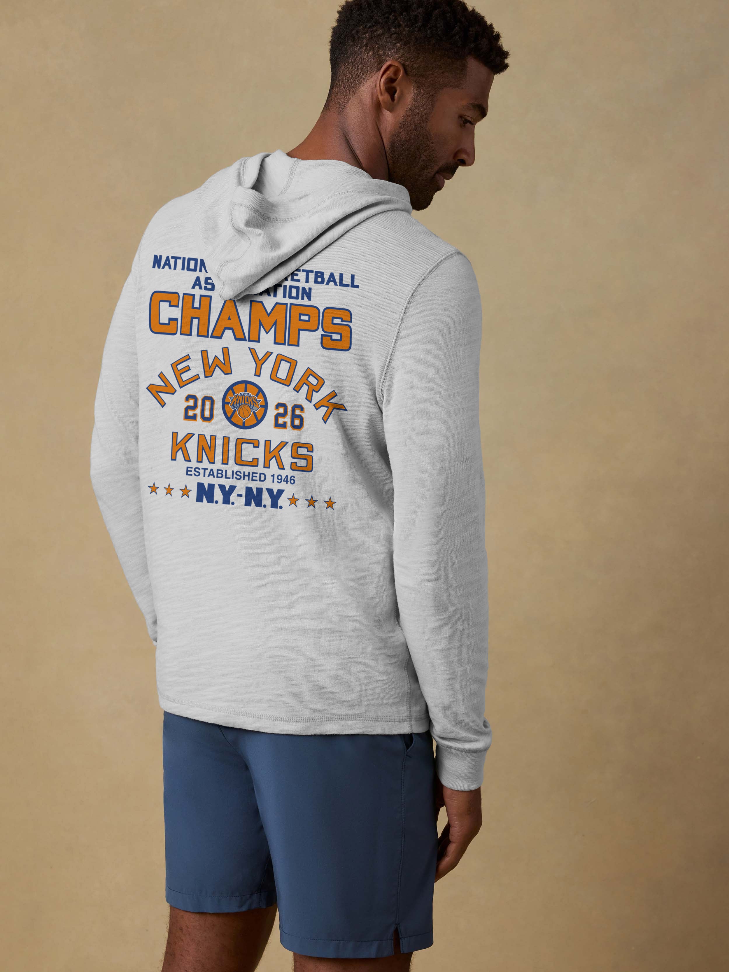 Man wearing a grey hoodie with a NY Knicks NBA Champs graphic on the back against a beige background