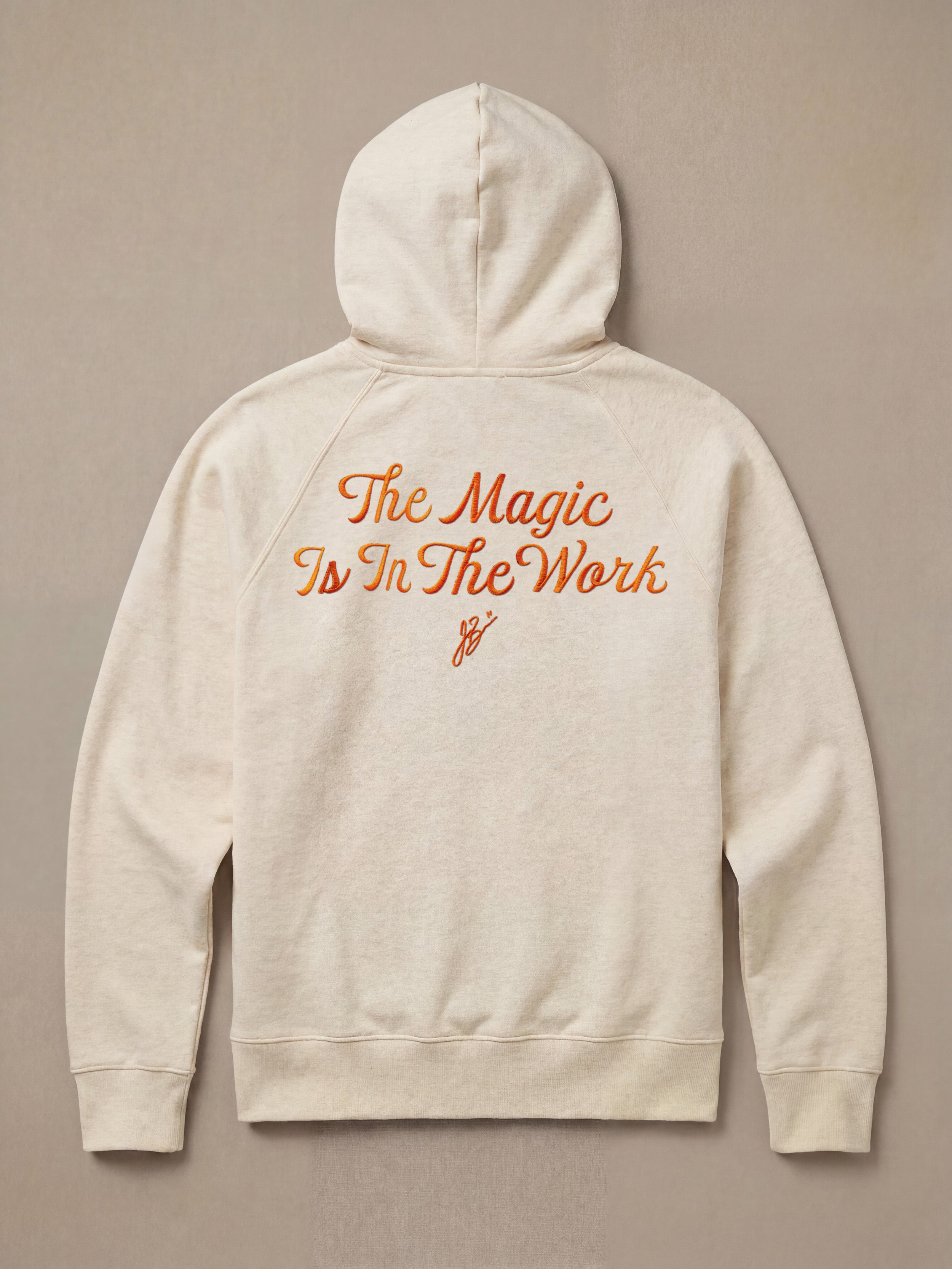 Heather hoodie with "The Magic Is In The Work" text on the back against a beige background
