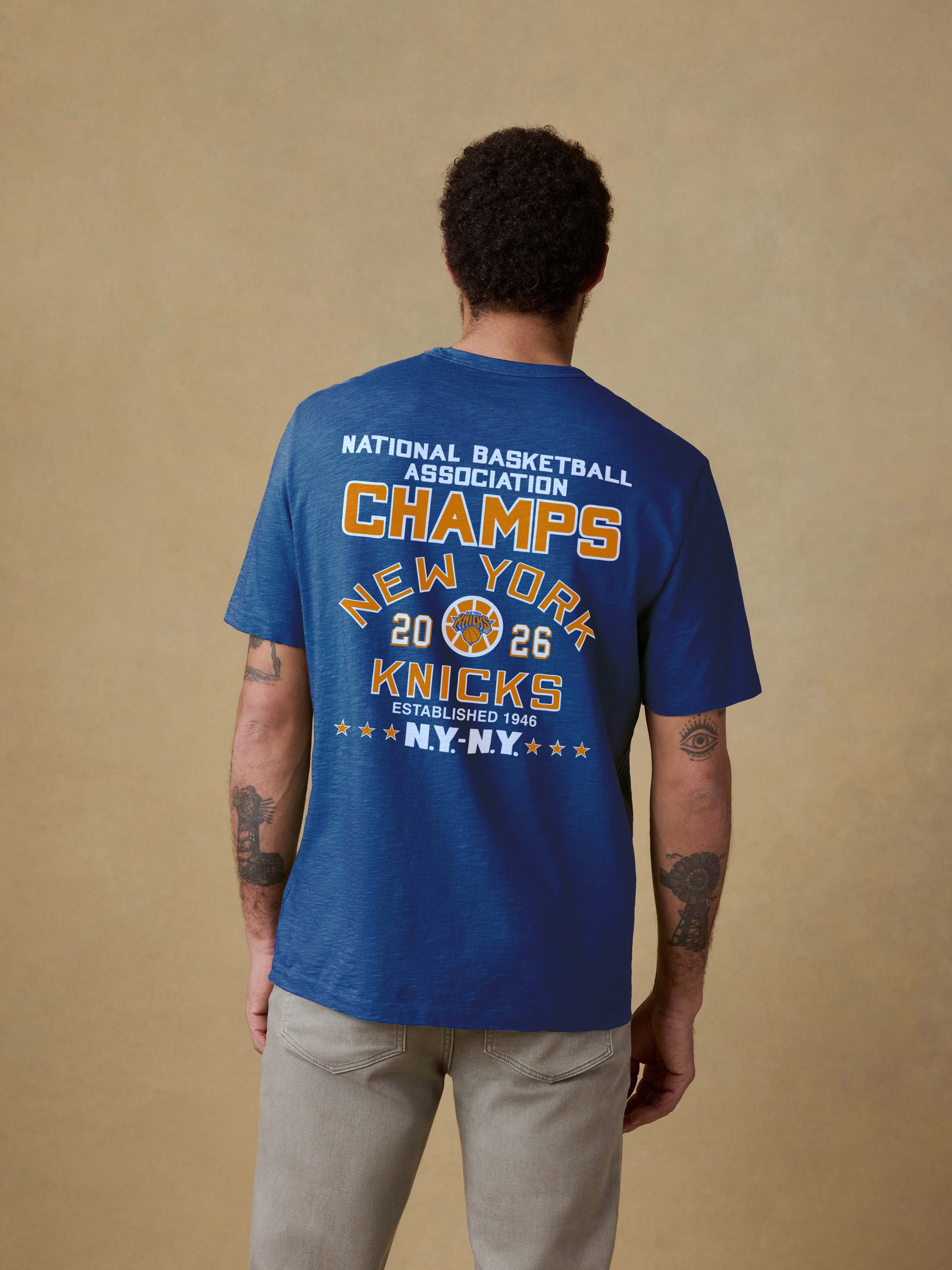 Man wearing a blue t-shirt with NY Knicks NBA Champs graphic on the back against a beige background