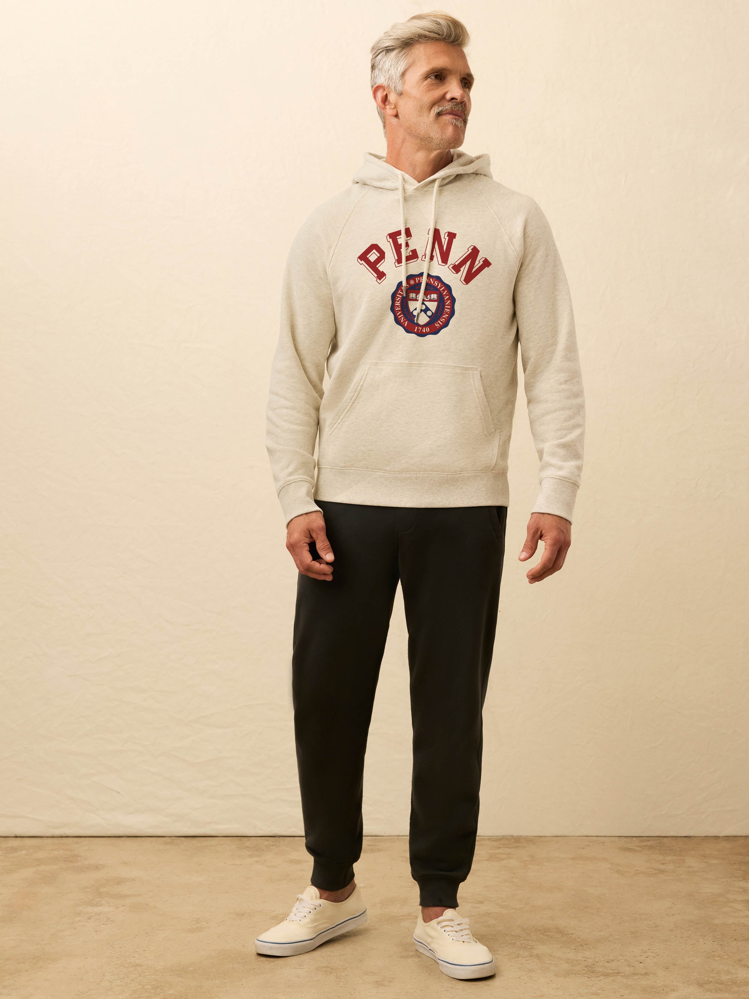 Man is wearing a heather grey hoodie with University of Pennsylvania graphic on the front against a beige background
