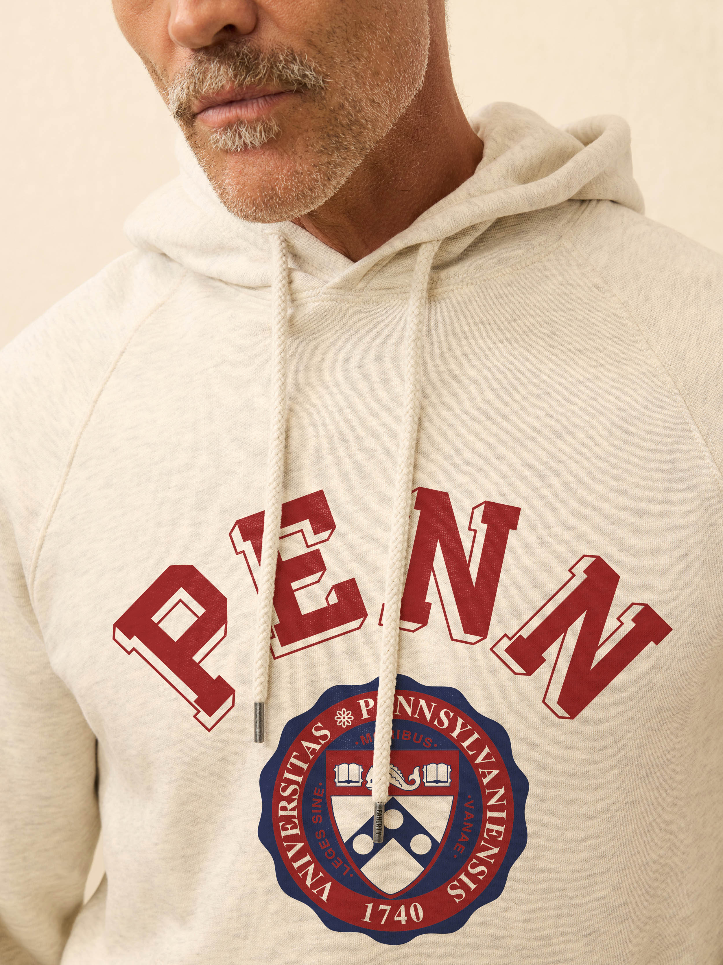 Man is wearing a heather grey hoodie with University of Pennsylvania graphic on the front against a beige background