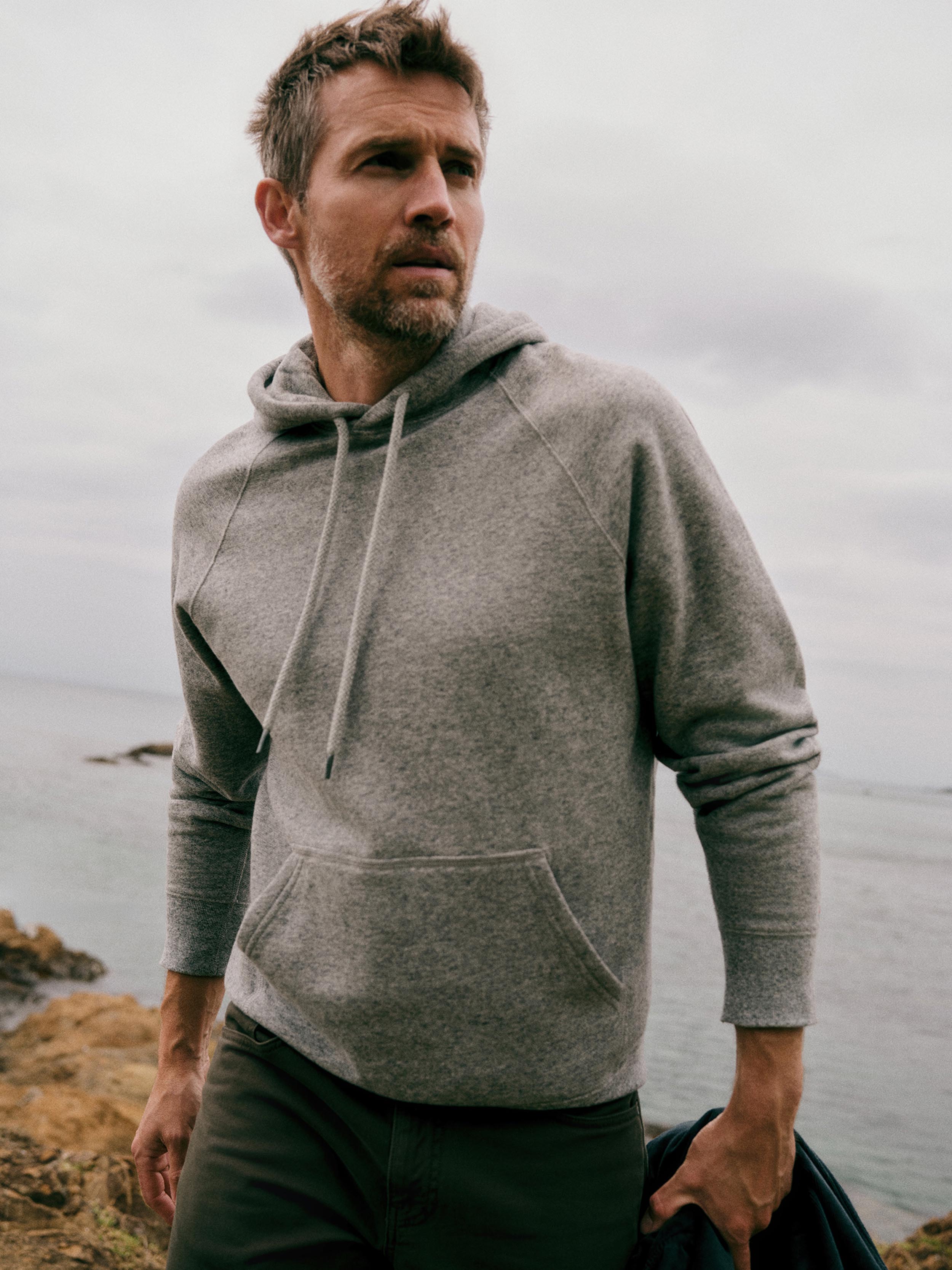 A man wearing a light gray heathered pullover hoodie with drawstrings and a kangaroo pocket, standing outdoors near the ocean under an overcast sky.