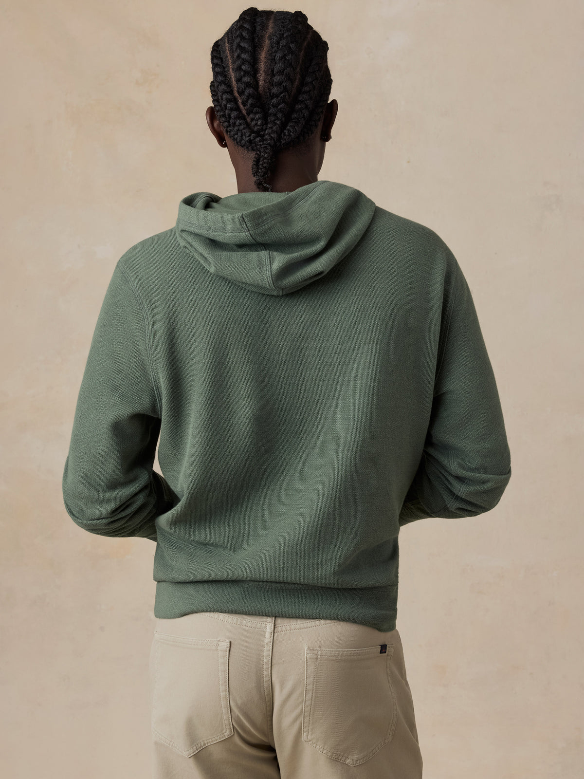 A man shown from the back wearing an olive green hooded sweatshirt and beige pants, standing against a textured beige wall., image 3 of 6