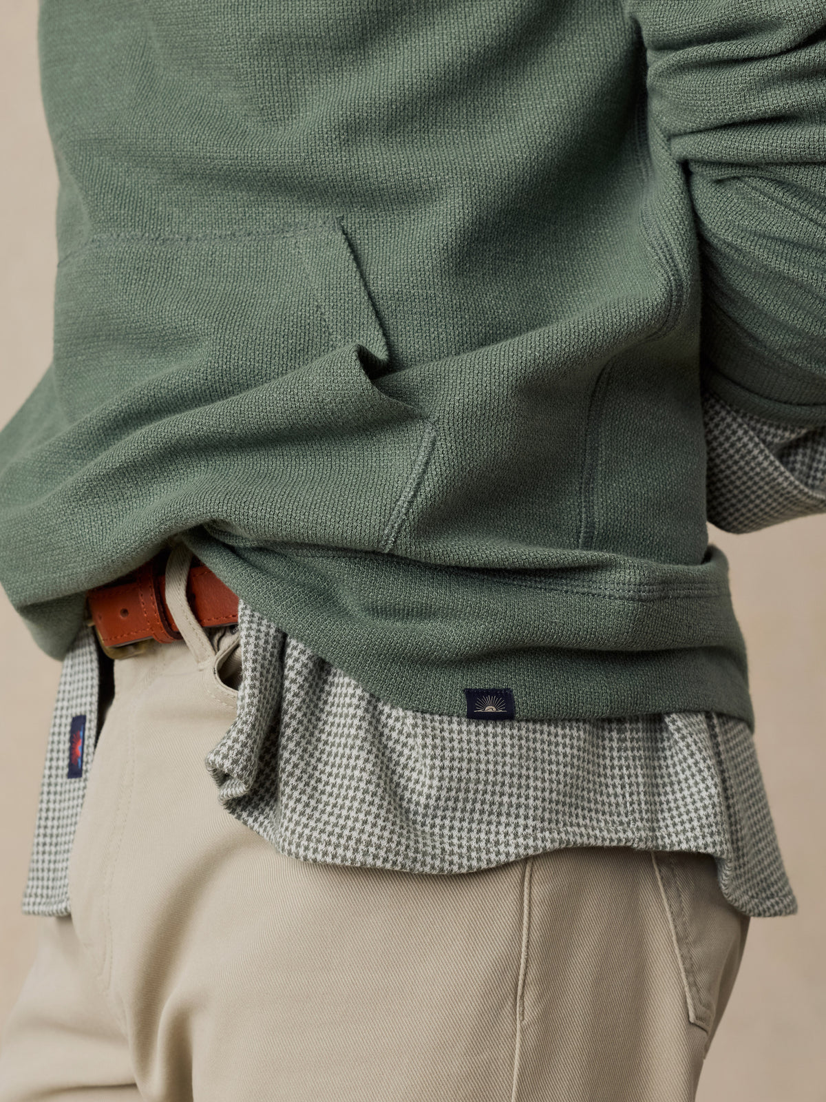 Close-up of a man wearing a sage green knit sweater layered over a small-scale checkered shirt and beige trousers, with a brown belt visible., image 4 of 6