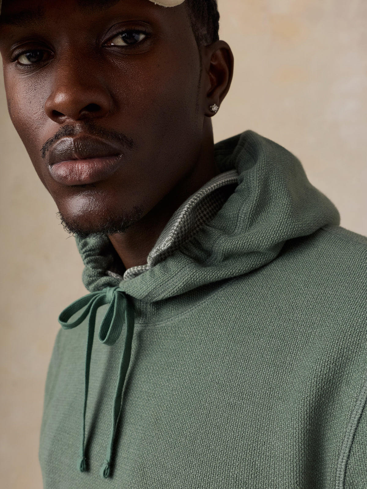 A close-up shot of a man wearing a sage green knit hoodie with a gray patterned inner hood lining and matching drawstrings, set against a warm beige background., image 6 of 6