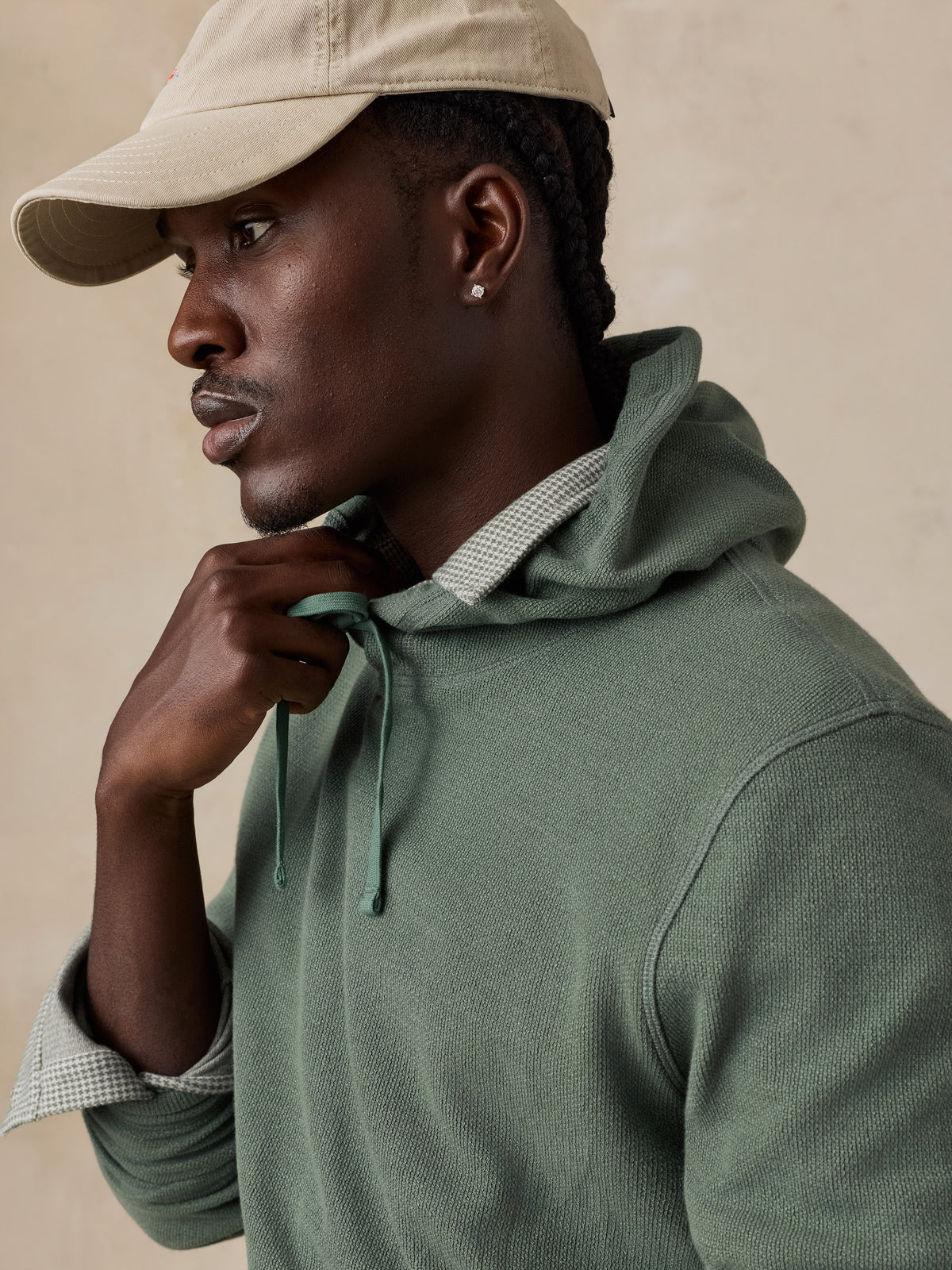 A man wearing an olive green knit hoodie with a matching drawstring, layered over a light-colored checkered shirt, and wearing a beige baseball cap, set against a warm beige background., image 2 of 6