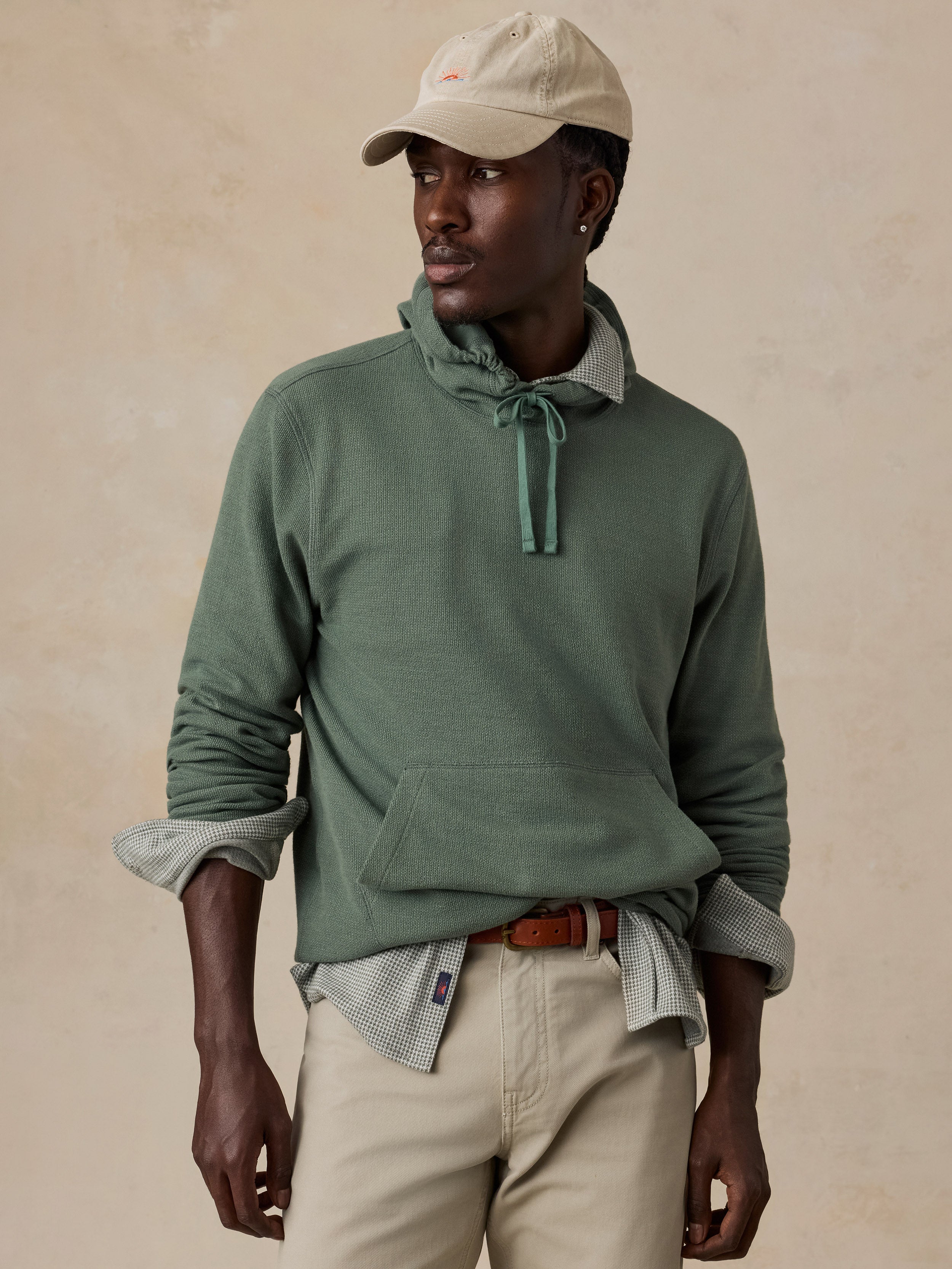 A man wearing a sage green hoodie layered over a light gray patterned shirt, paired with beige pants and a beige baseball cap, against a textured beige background.