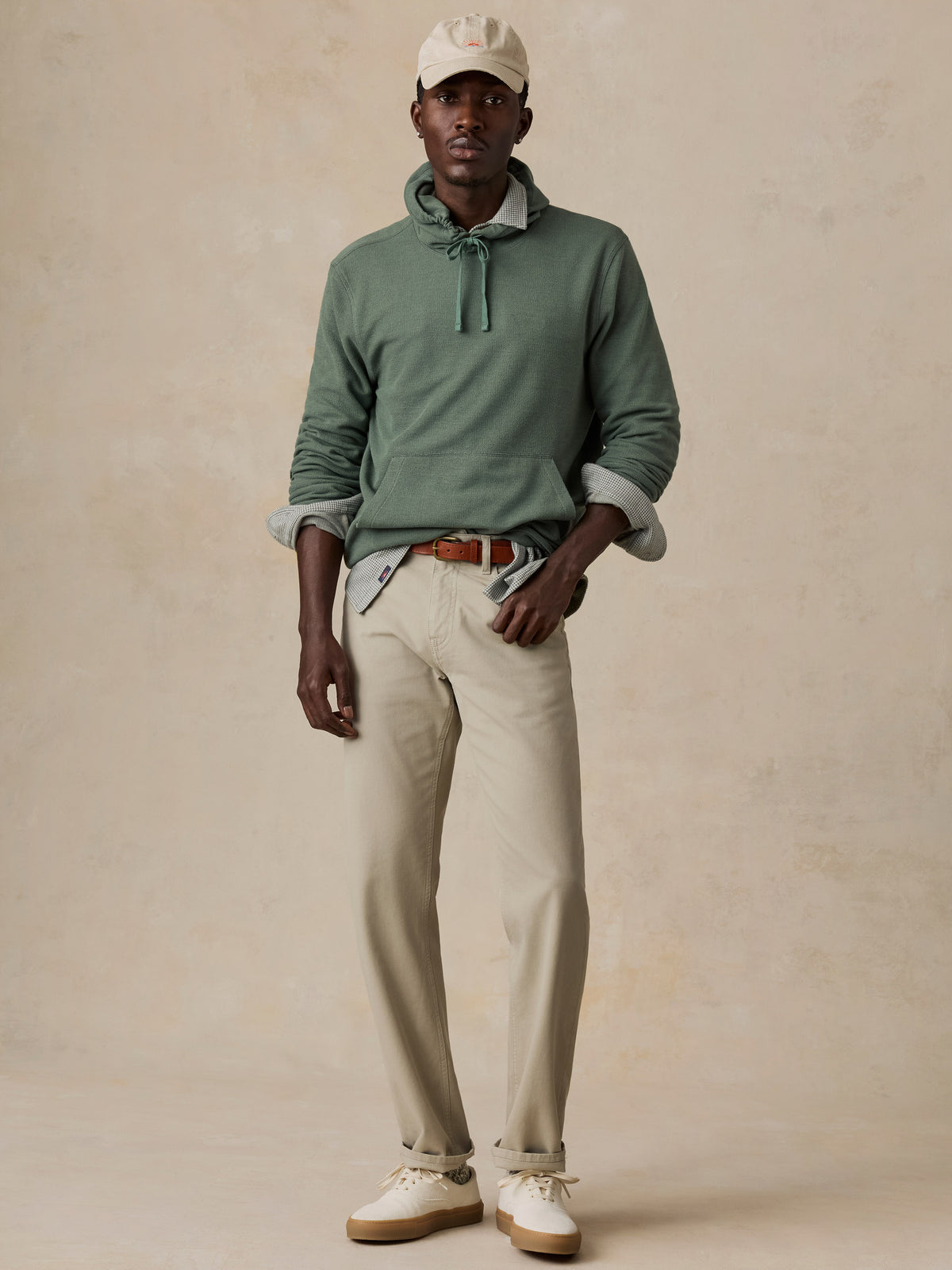 A man modeling a sage green knit pullover sweater with a tie-neck detail, styled with beige trousers, a brown leather belt, a light green shirt underneath, a cream cap, and white sneakers against a textured beige studio backdrop., image 5 of 6