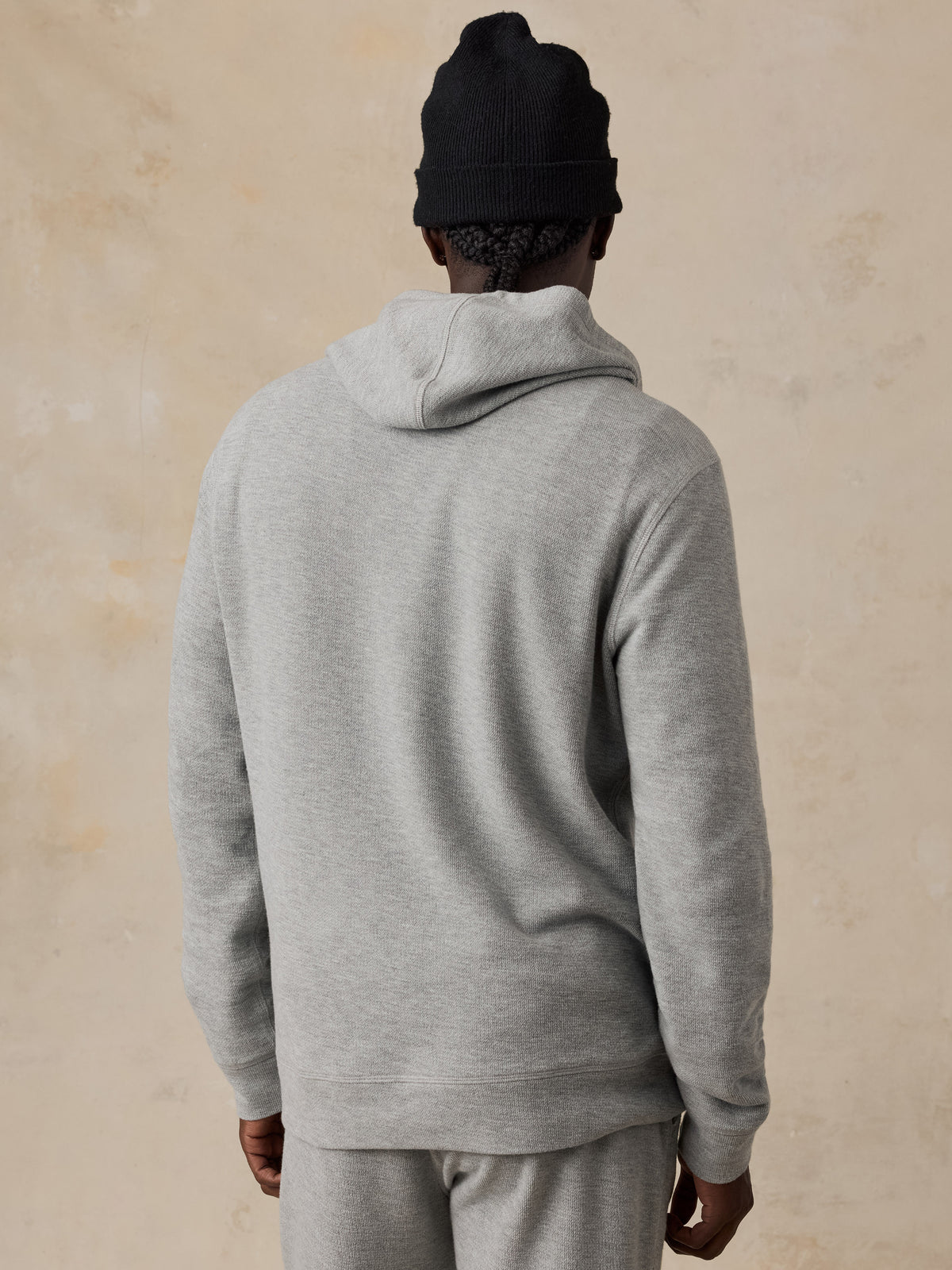 A man shown from the back wearing a light heather gray hooded sweatshirt and matching gray sweatpants, with a black knit beanie, against a warm beige textured wall., image 4 of 6