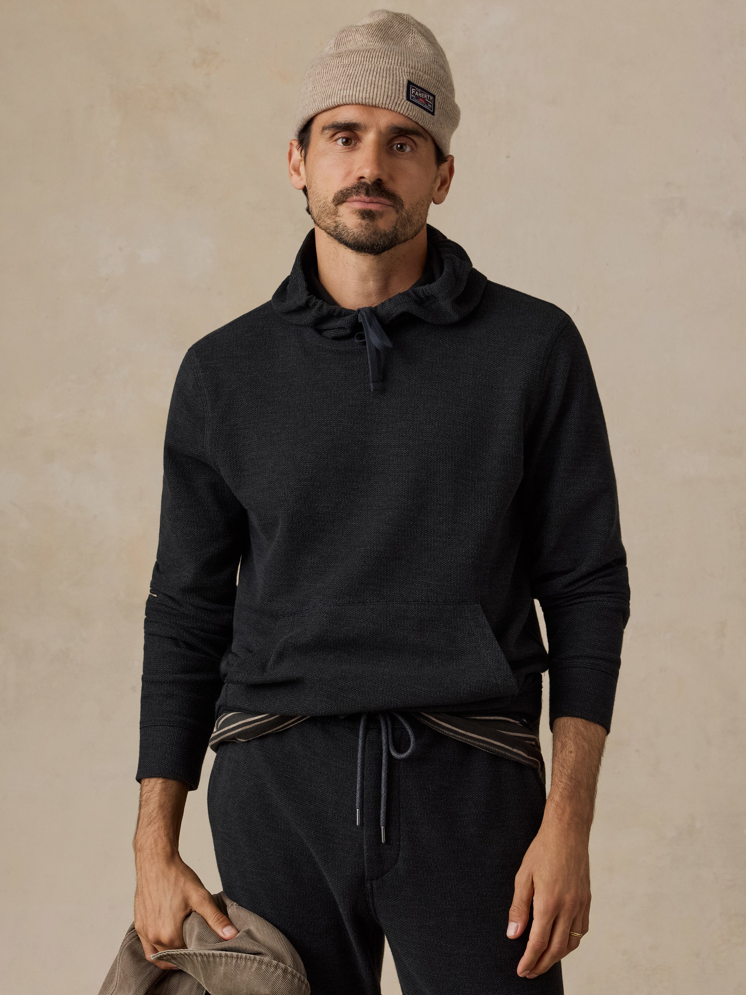 A man wearing a dark charcoal gray knit hoodie with a drawstring neck, matching dark sweatpants, a beige beanie, and holding a light beige jacket, set against a textured beige wall.
