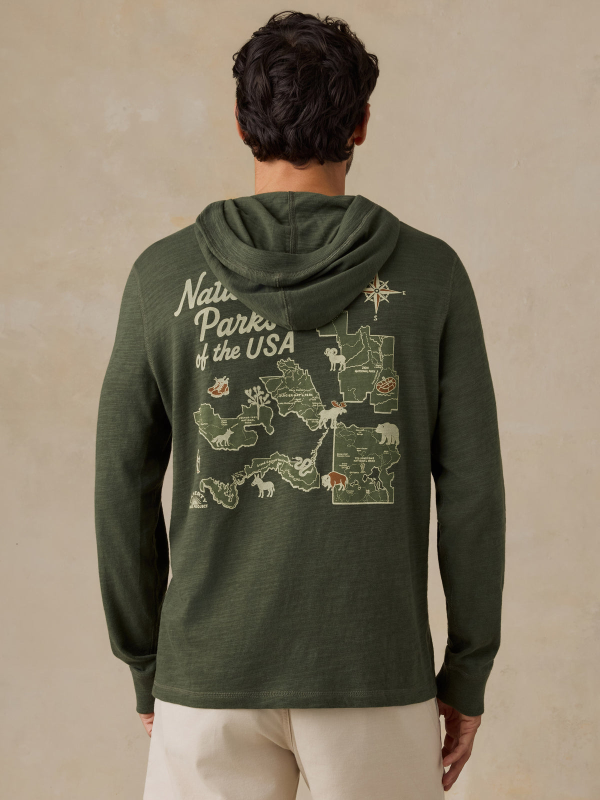 A man viewed from behind wearing an olive green hooded sweatshirt with a large 'National Parks of the USA' map graphic printed on the back, paired with light beige shorts, against a textured beige background., image 4 of 8