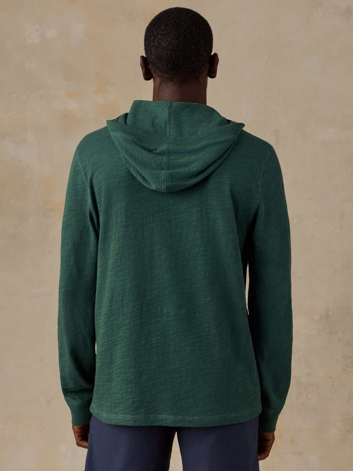 A man viewed from behind wearing a dark green hooded long-sleeve shirt and navy pants, standing against a textured beige wall., image 3 of 5