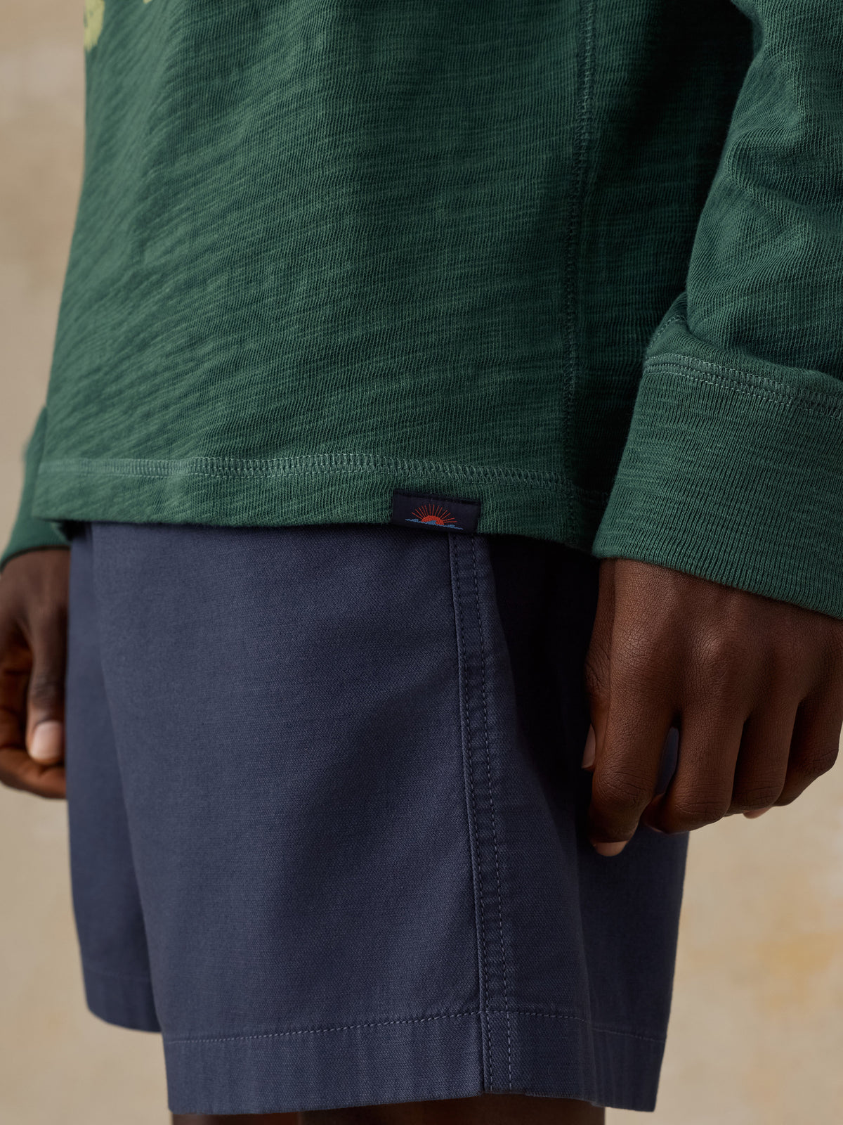 Close-up of a man wearing a dark green long-sleeve hoodie and navy blue shorts, with a small Faherty brand sun and waves tag visible on the hem of the hoodie., image 5 of 5