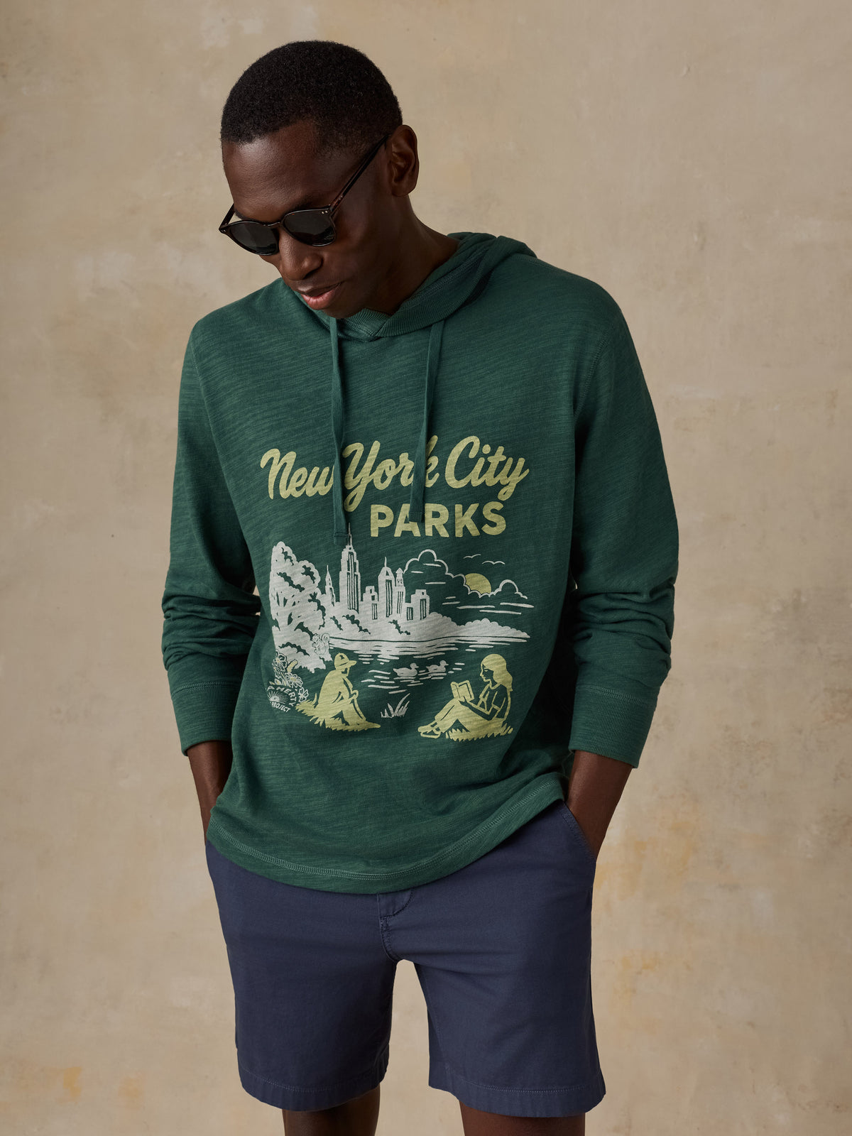 A man wearing a dark green heathered hoodie with a vintage-style 'New York City Parks' graphic on the front, paired with navy blue shorts, set against a textured beige background., image 4 of 5