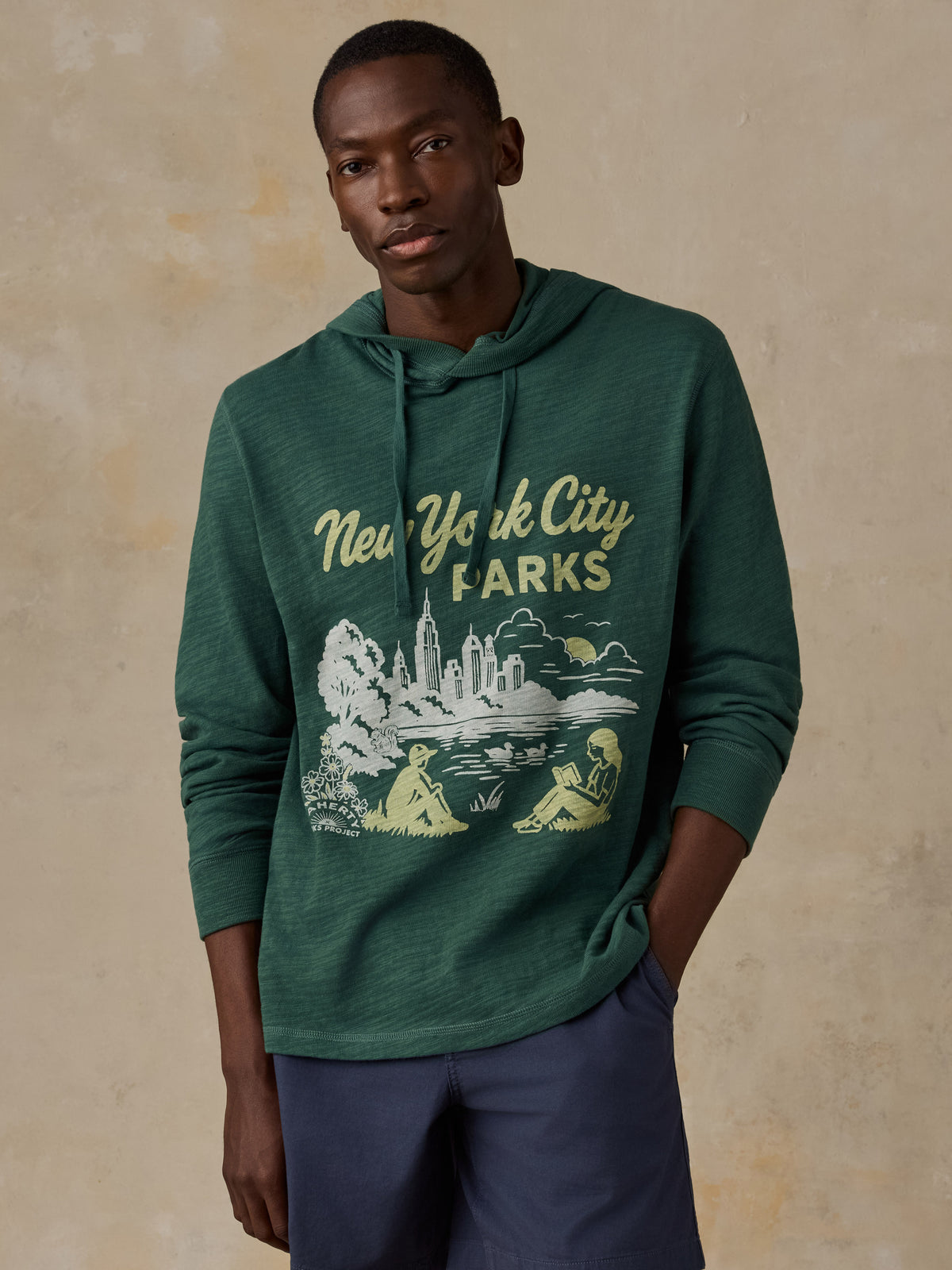 A man wearing a dark green heathered pullover hoodie with a 'New York City Parks' graphic on the front, paired with navy blue pants, standing against a textured beige background., image 1 of 5