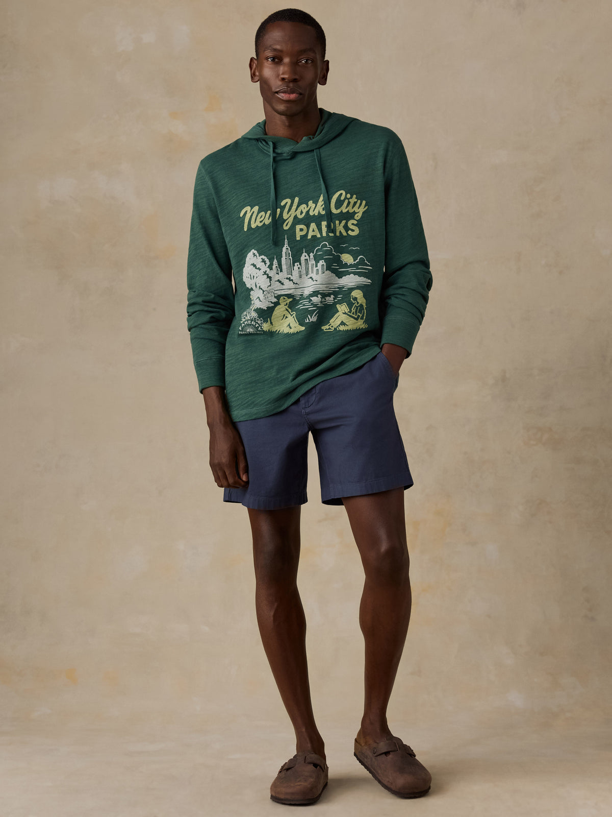 A man modeling a dark green pullover hoodie with a 'New York City Parks' graphic on the front, paired with navy blue shorts and brown slip-on shoes, against a textured beige studio backdrop., image 2 of 5