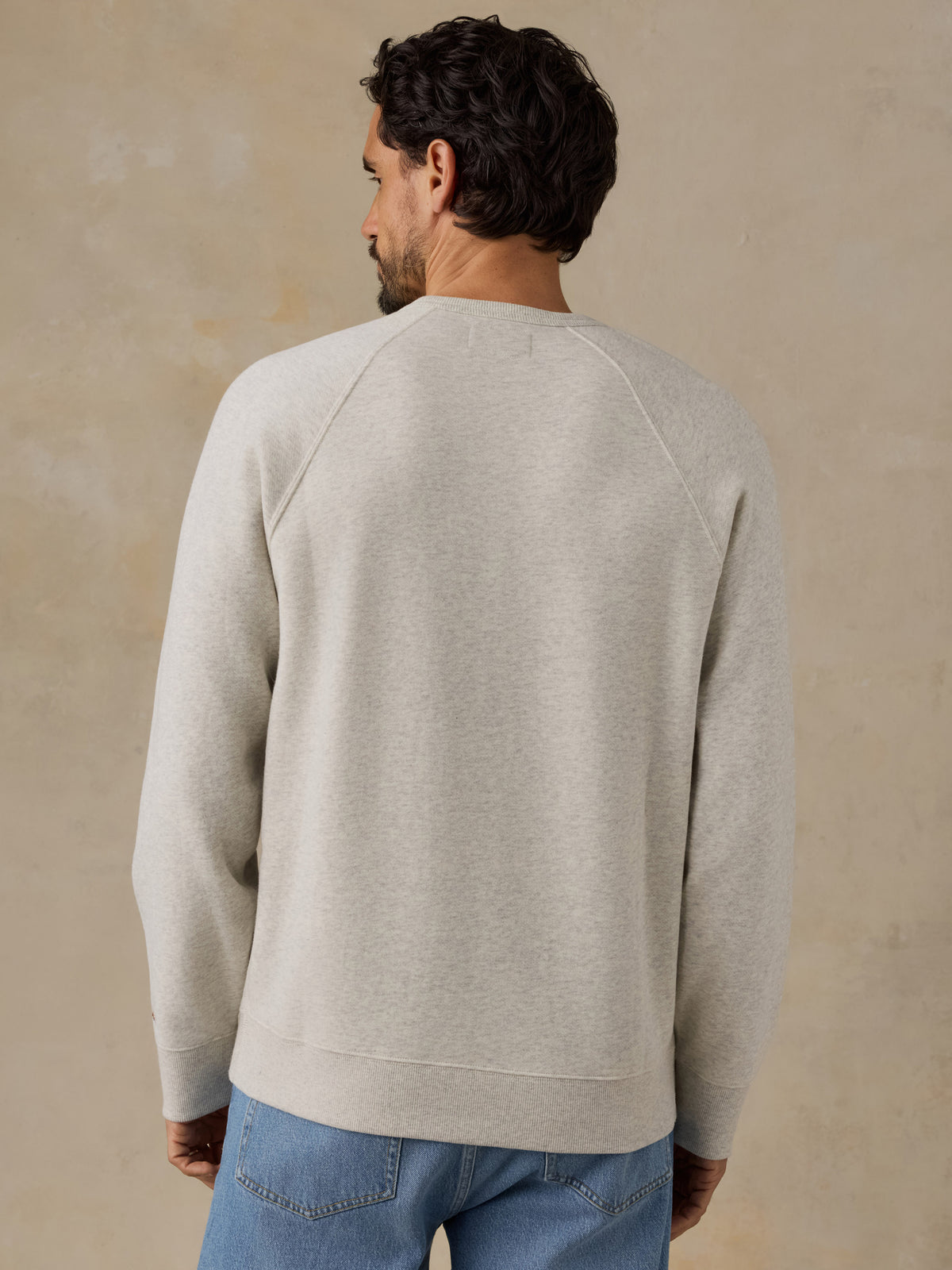 A man viewed from behind wearing a light beige crewneck sweatshirt and blue jeans, standing against a textured beige wall., image 3 of 7