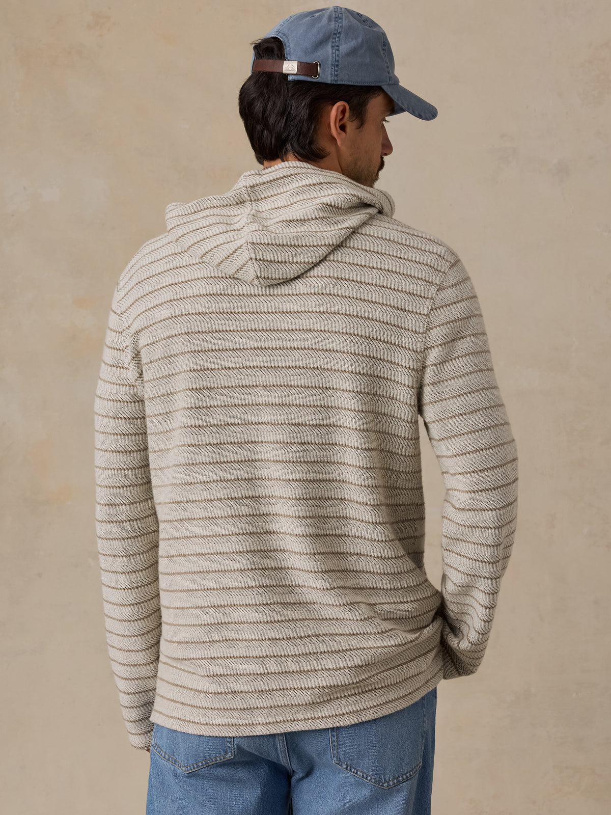 A man wearing a beige and gray horizontally striped knit hoodie, blue jeans, and a blue baseball cap, shown from a back-three-quarter view against a textured beige wall., image 3 of 6