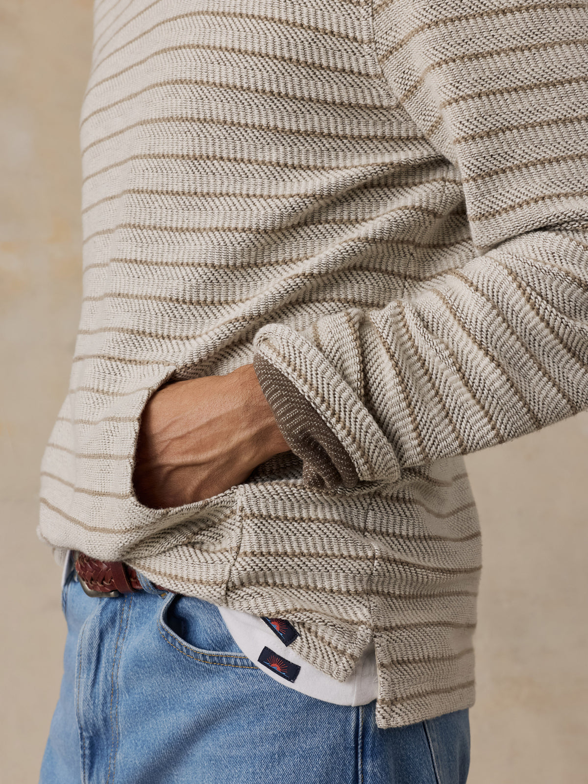 Close-up of a man wearing a beige and brown horizontally striped ribbed knit sweater with a pocket, tucked into blue jeans., image 4 of 6