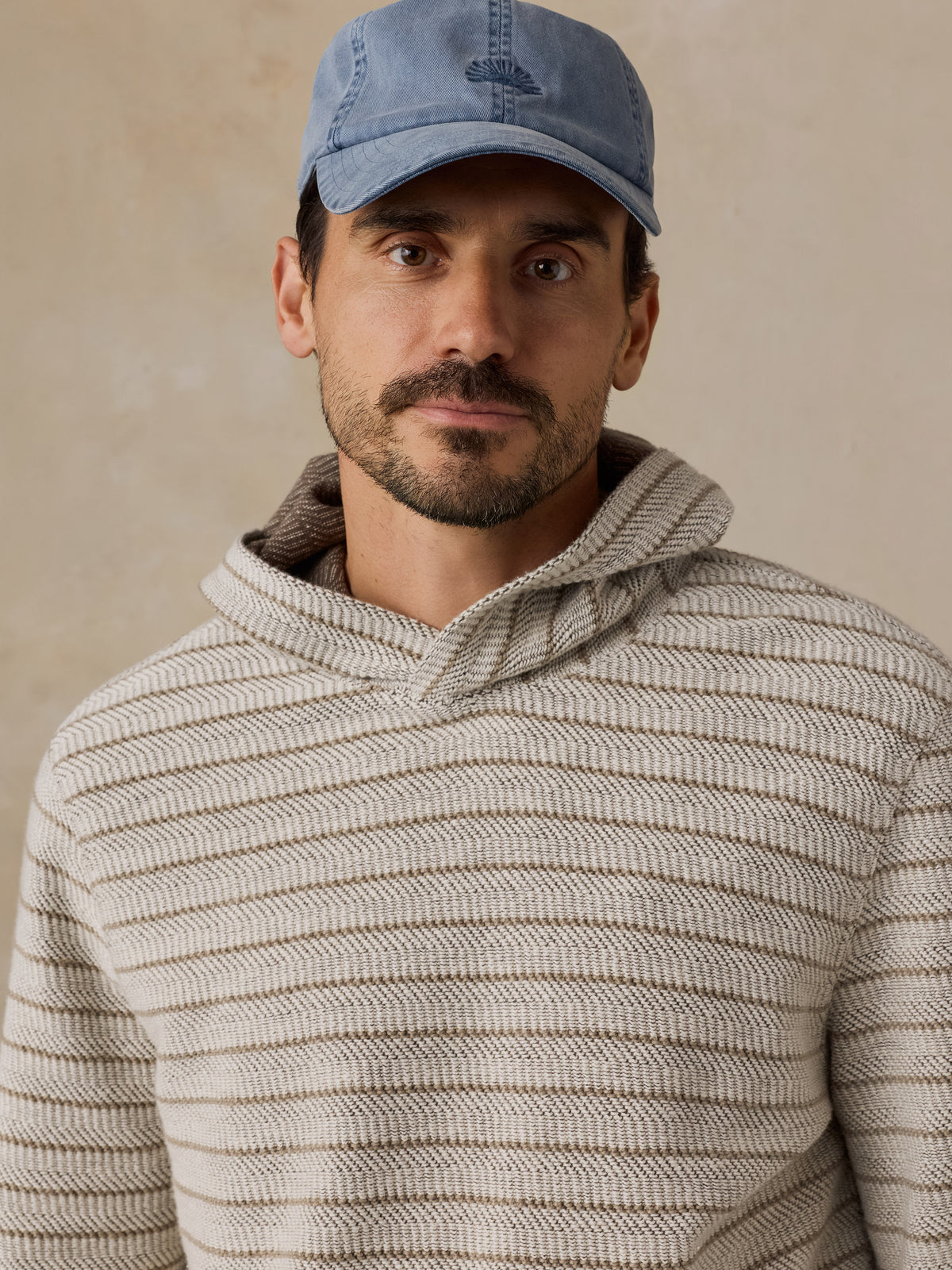 A man wearing a light blue baseball cap and a beige textured knit hoodie, photographed against a neutral beige background., image 6 of 6