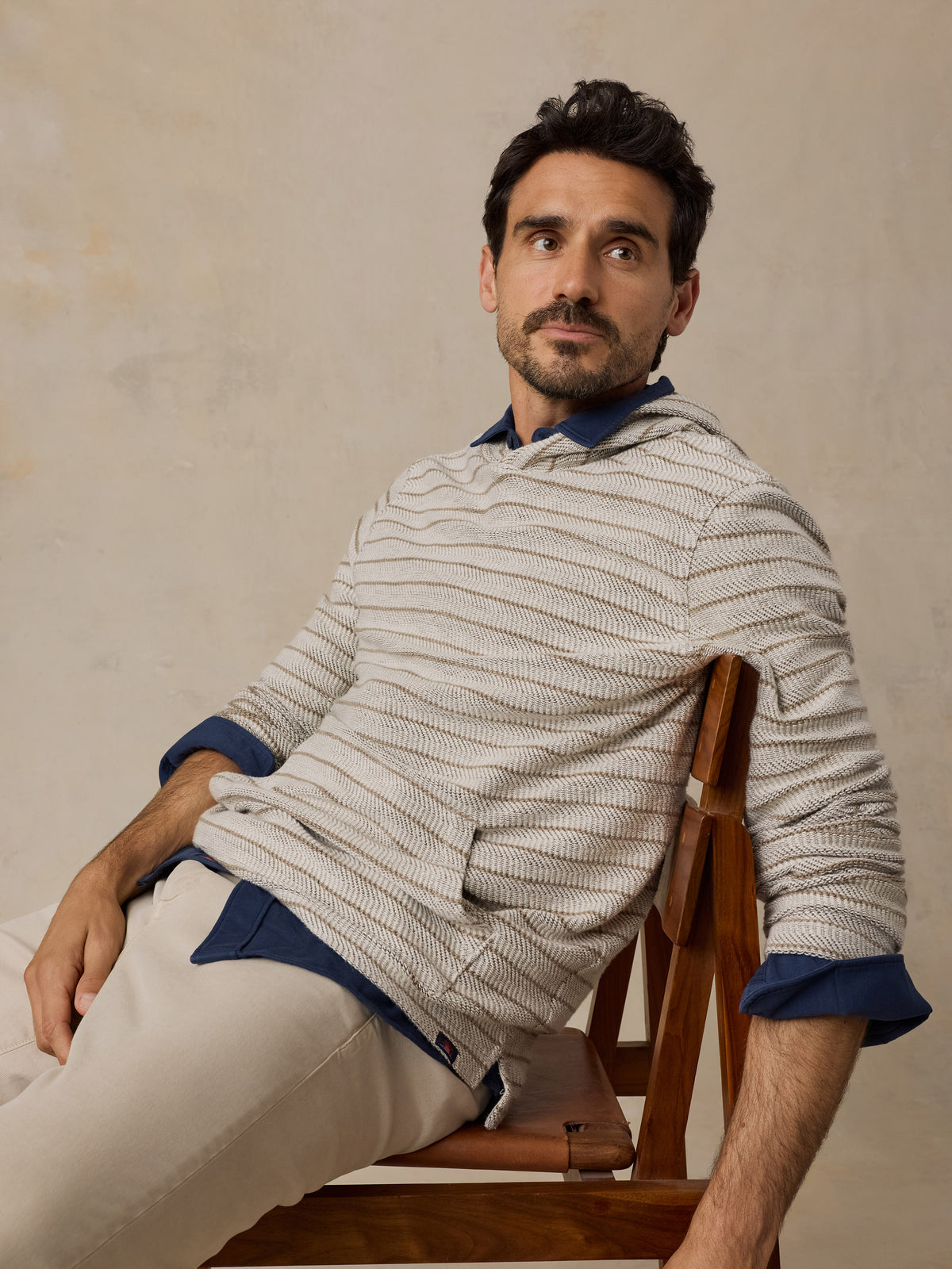 A man wearing a beige and white striped knit sweater over a blue collared shirt, paired with beige trousers, sitting on a wooden chair against a textured beige wall., image 2 of 6