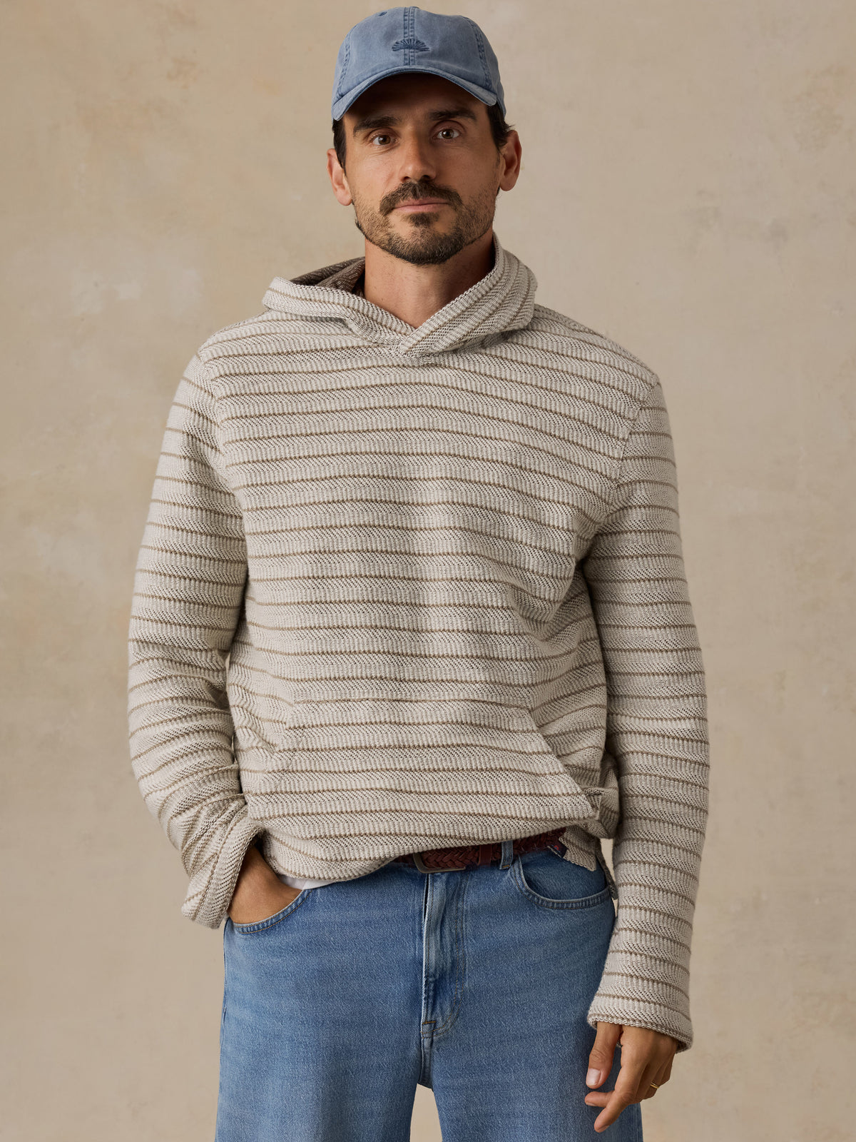A man wearing a beige and gray horizontally striped knit hoodie with a cowl neck, paired with blue jeans and a blue baseball cap, standing against a textured beige wall., image 1 of 6