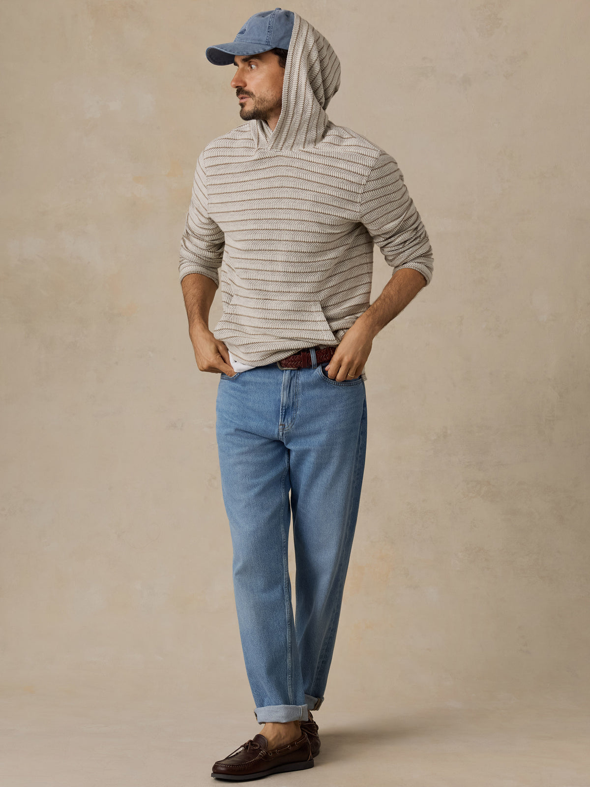 A man wearing a beige and gray striped hoodie, blue jeans, a blue baseball cap, and brown leather loafers, standing against a textured beige studio backdrop., image 5 of 6