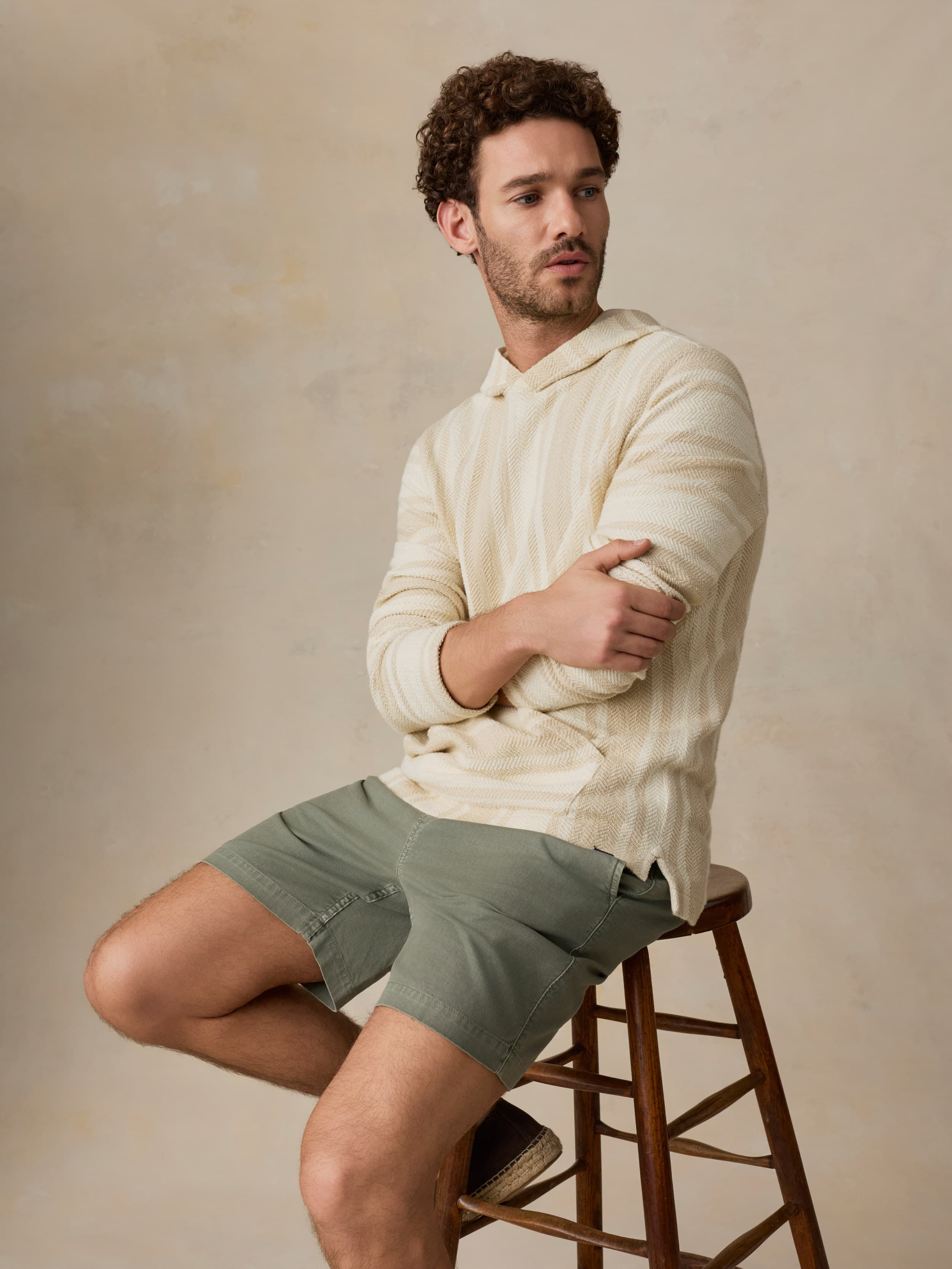 Man sitting on a wooden stool wearing a cream hoodie and green shorts against a beige background
