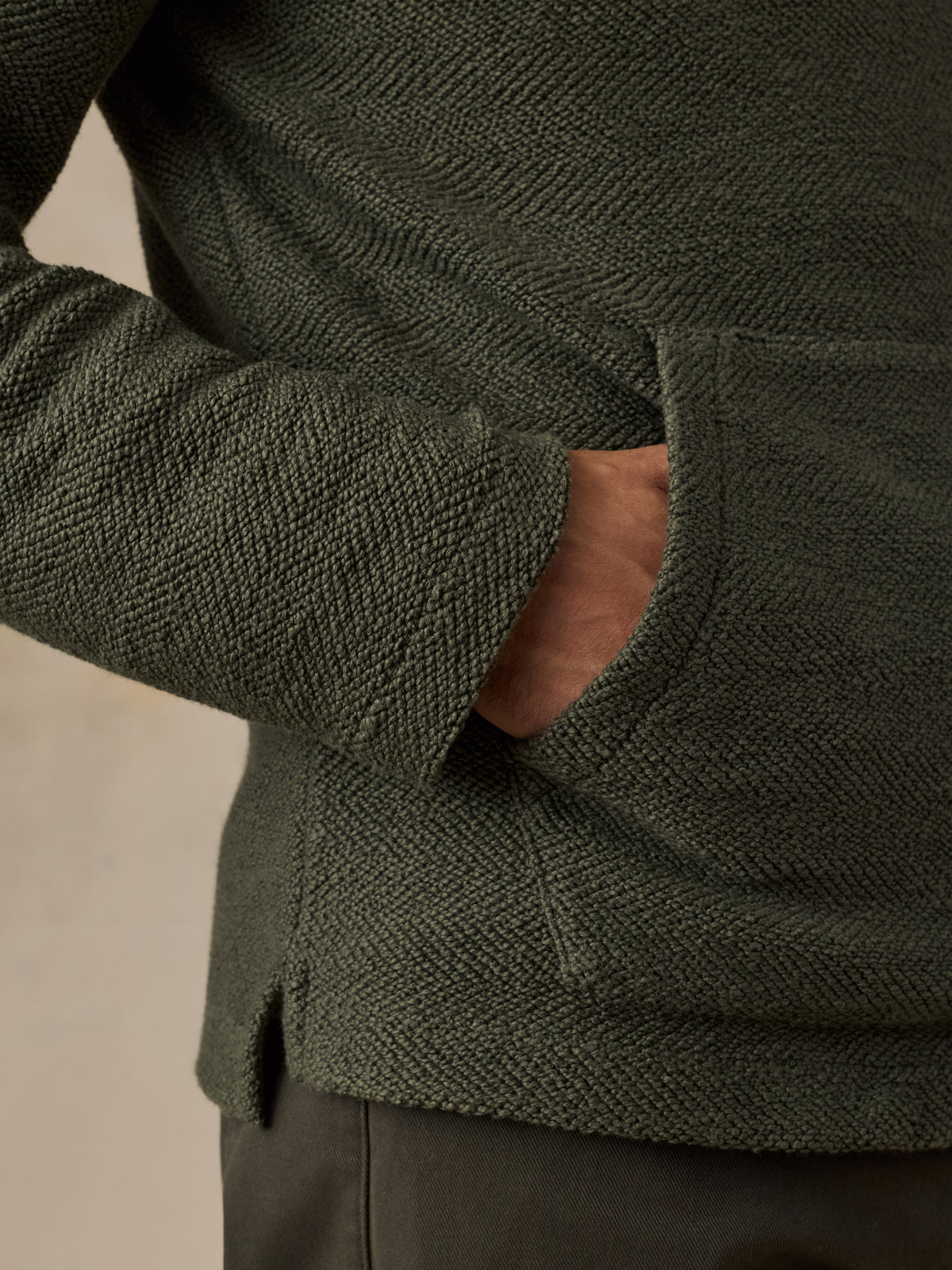 Close-up of a man wearing a dark olive green textured knit sweater with a side pocket, showing the fabric weave and a hand in the pocket., image 4 of 6