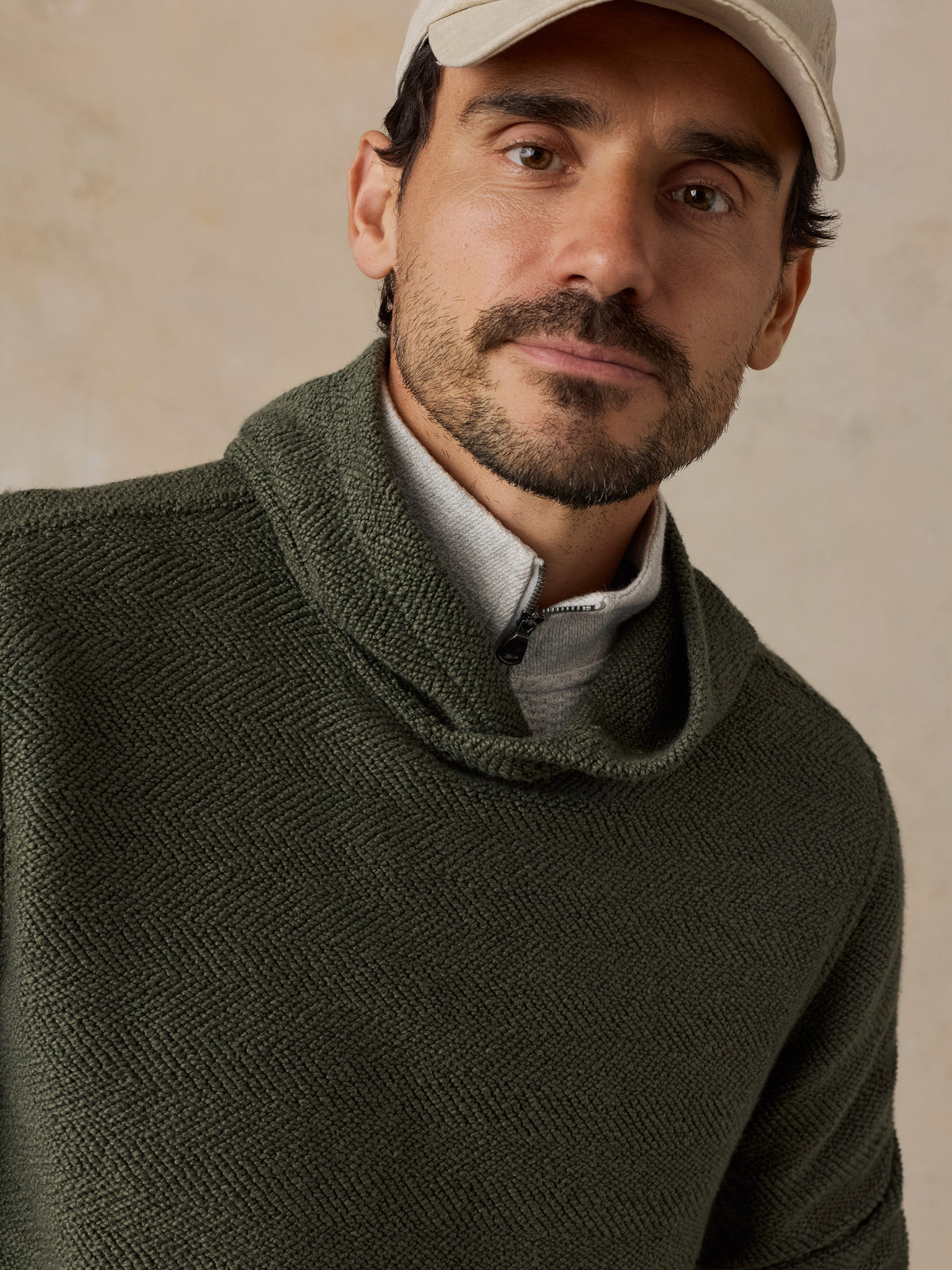 A close-up of a man wearing an olive green textured knit cowl-neck sweater over a light gray collared shirt, with a beige baseball cap, against a neutral beige background., image 5 of 6