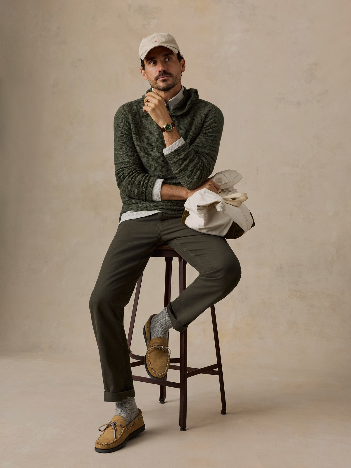 A man sits on a dark wooden stool wearing an olive green knit sweater, matching olive trousers, tan boat shoes, gray socks, and a beige baseball cap. He holds a white canvas tote bag against a textured beige studio backdrop., image 6 of 6