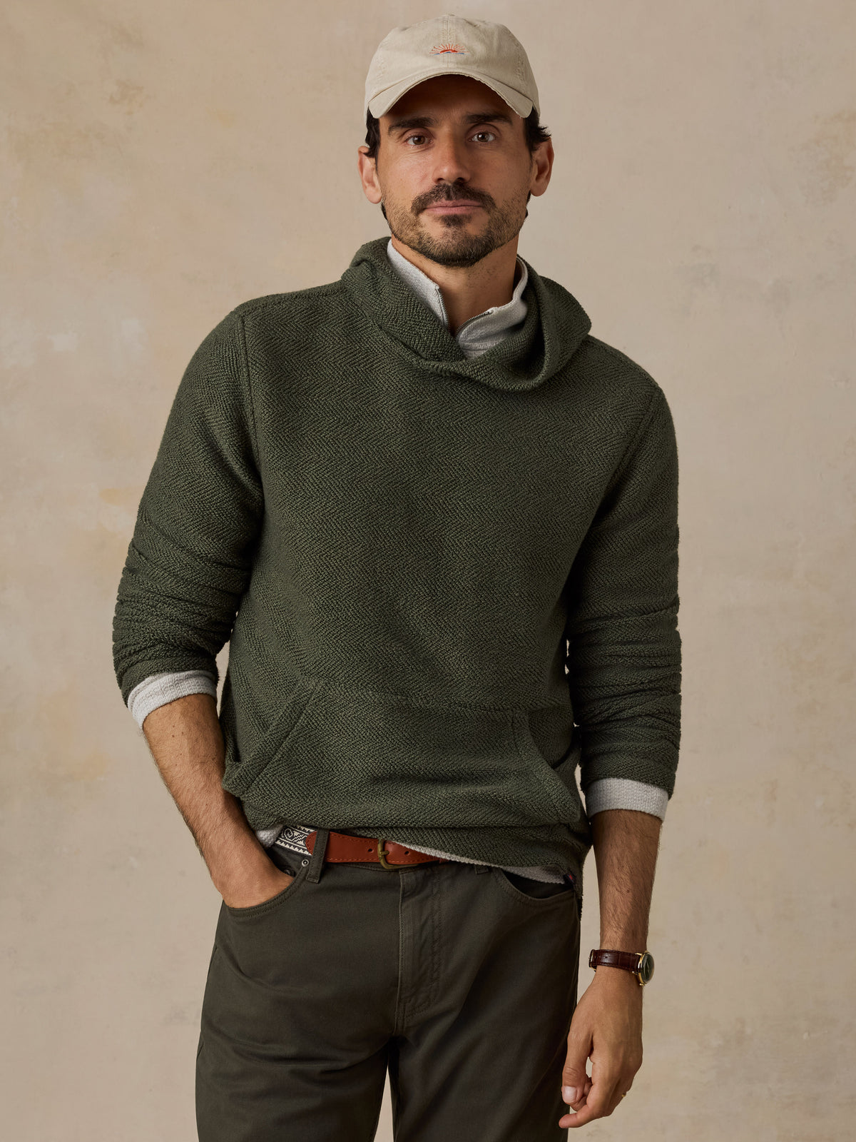 A man wearing an olive green cowl-neck knit sweater over a light gray collared shirt, paired with dark olive pants, a beige baseball cap, and a brown belt, standing against a textured beige wall., image 1 of 6