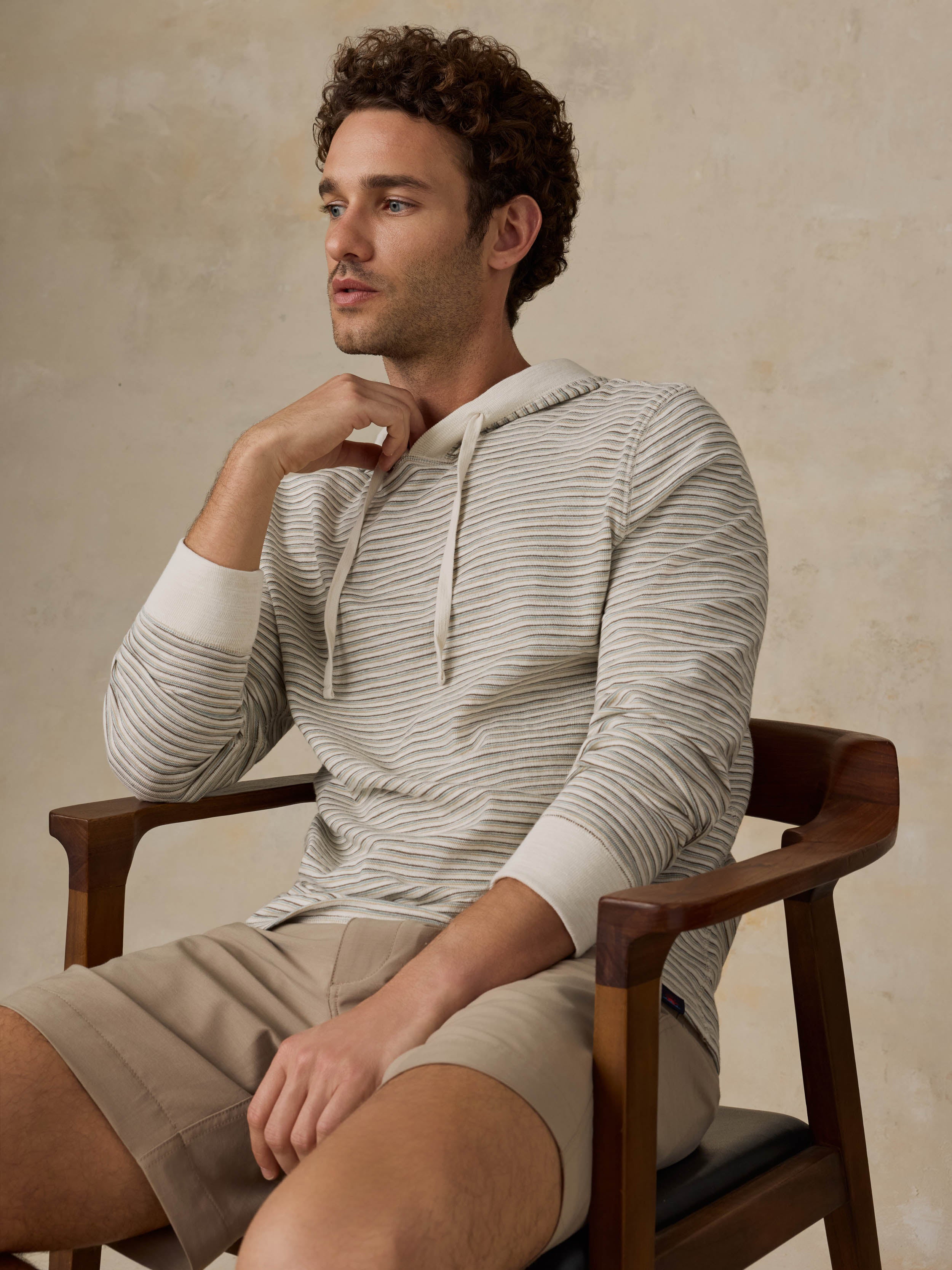 A man wearing a light beige and gray striped hooded sweater and tan shorts, standing against a textured beige wall.