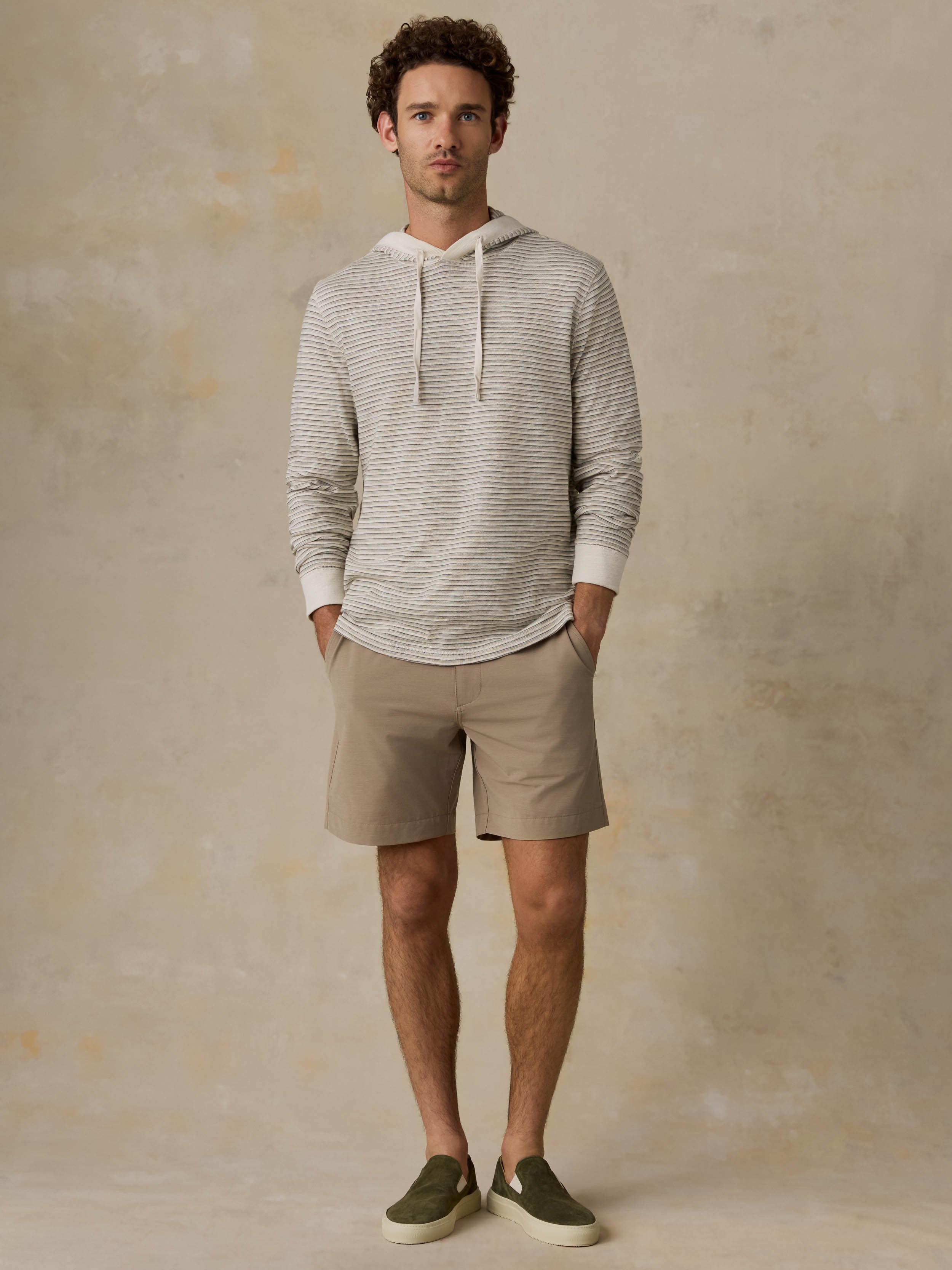 A man wearing a light beige and gray striped hooded sweater and tan shorts, standing against a textured beige wall.
