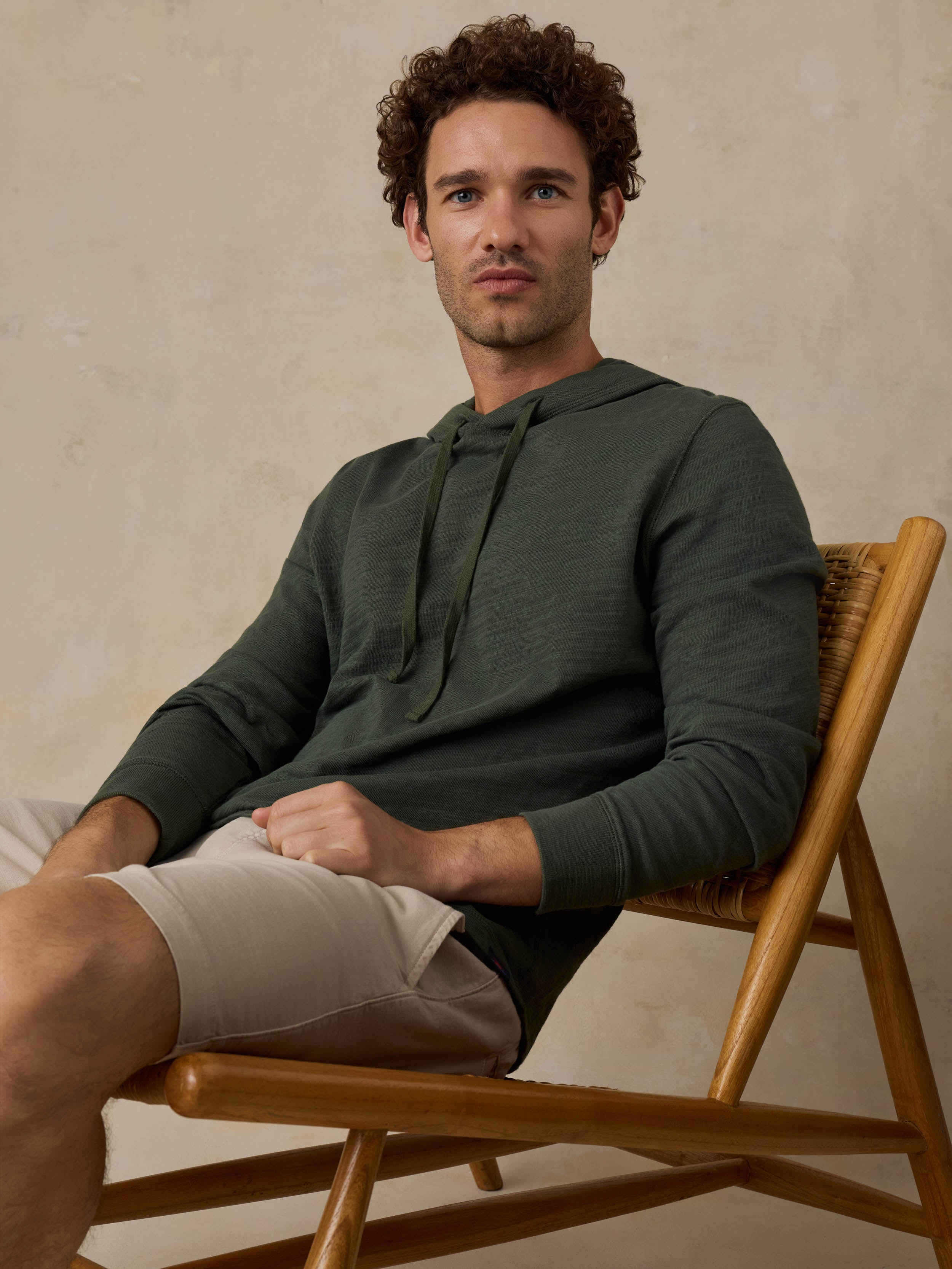 Man sitting on a wooden chair wearing a dark olive green hoodie and beige shorts against a beige background