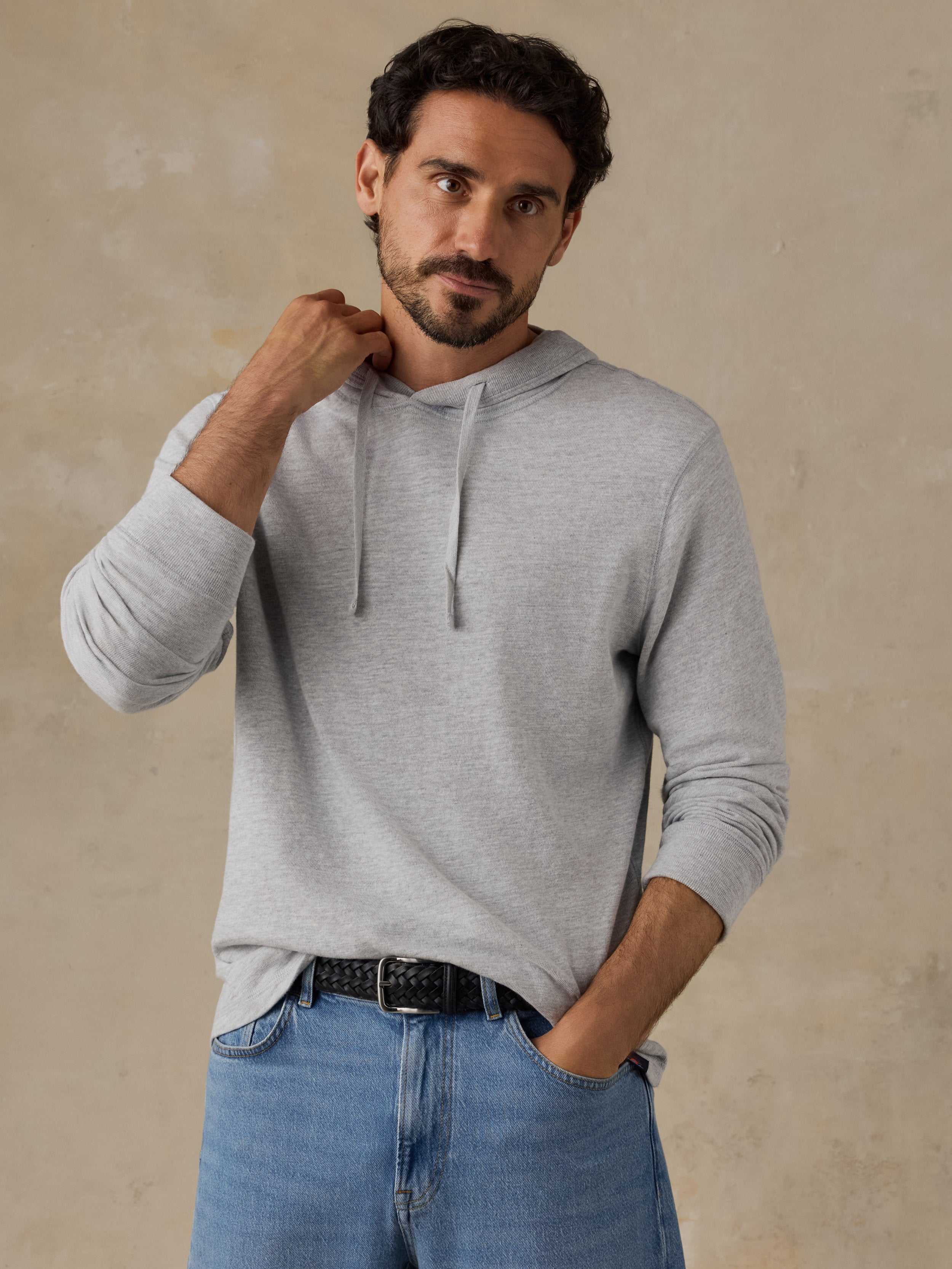 A man wearing a light gray long-sleeve hoodie with drawstrings, paired with blue jeans and a black belt, standing against a textured beige wall.