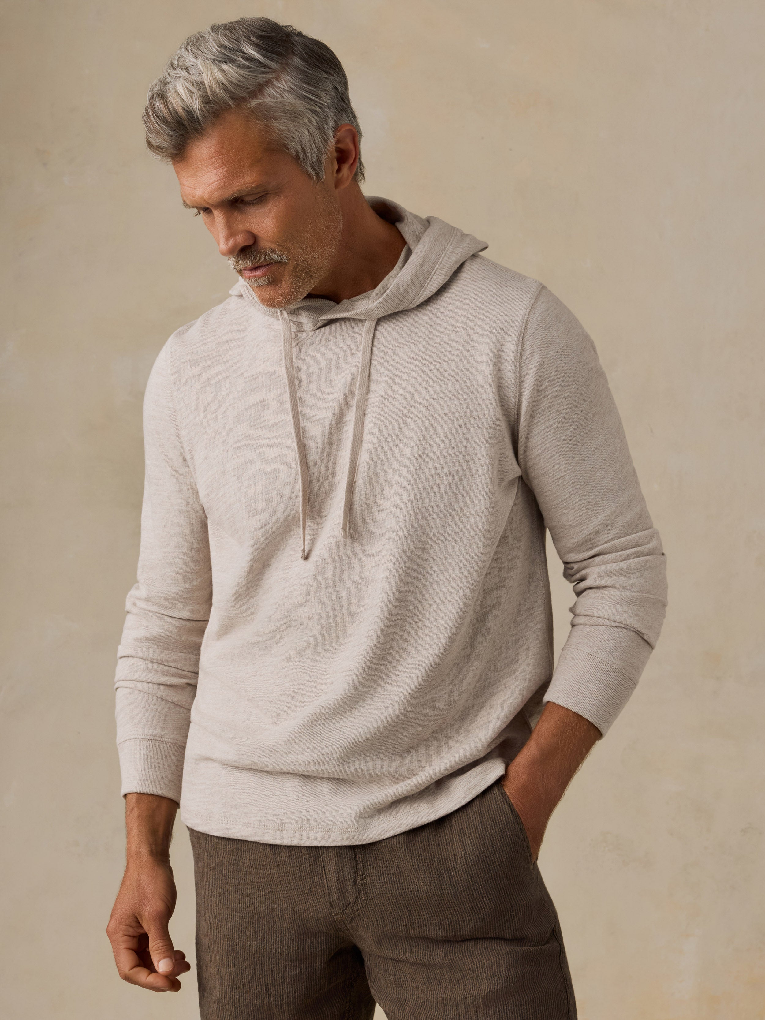 A man wearing a light beige long-sleeve knit hoodie with drawstrings and brown trousers, standing against a textured beige background.