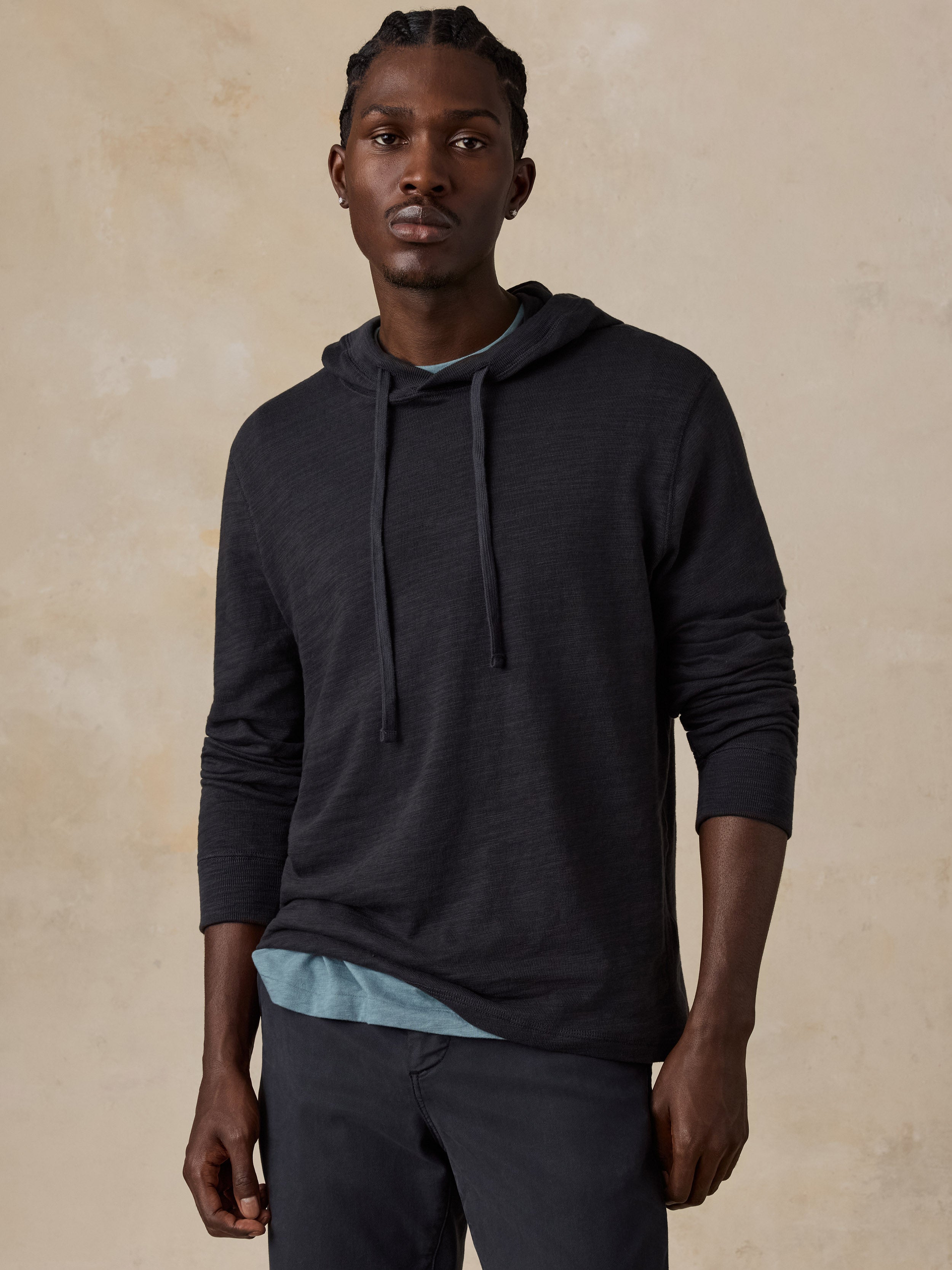 Man wearing a black hoodie over a blue t-shirt and navy chino pants against a beige studio background