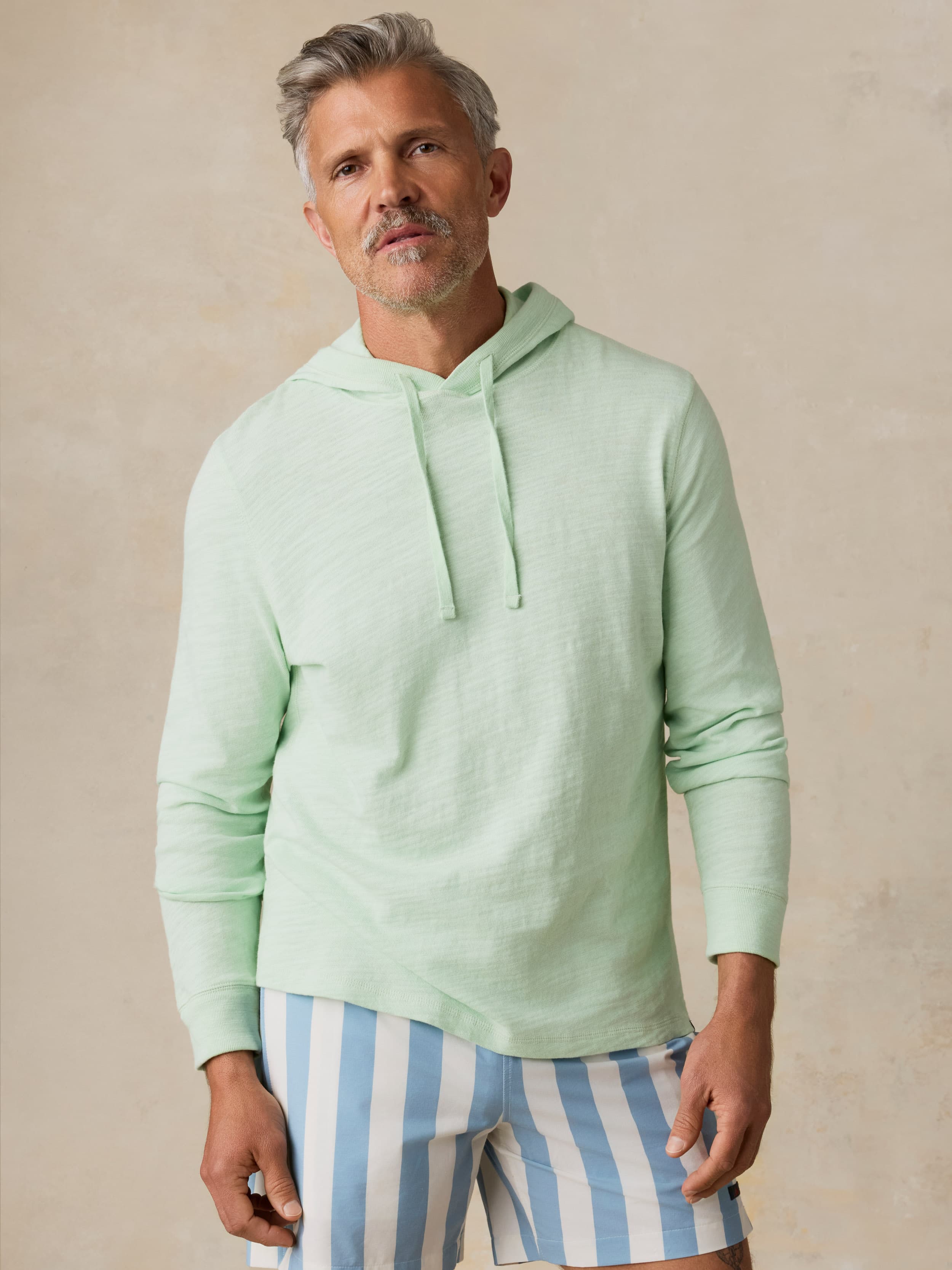 Man wearing a light green hoodie and blue striped shorts against a beige background