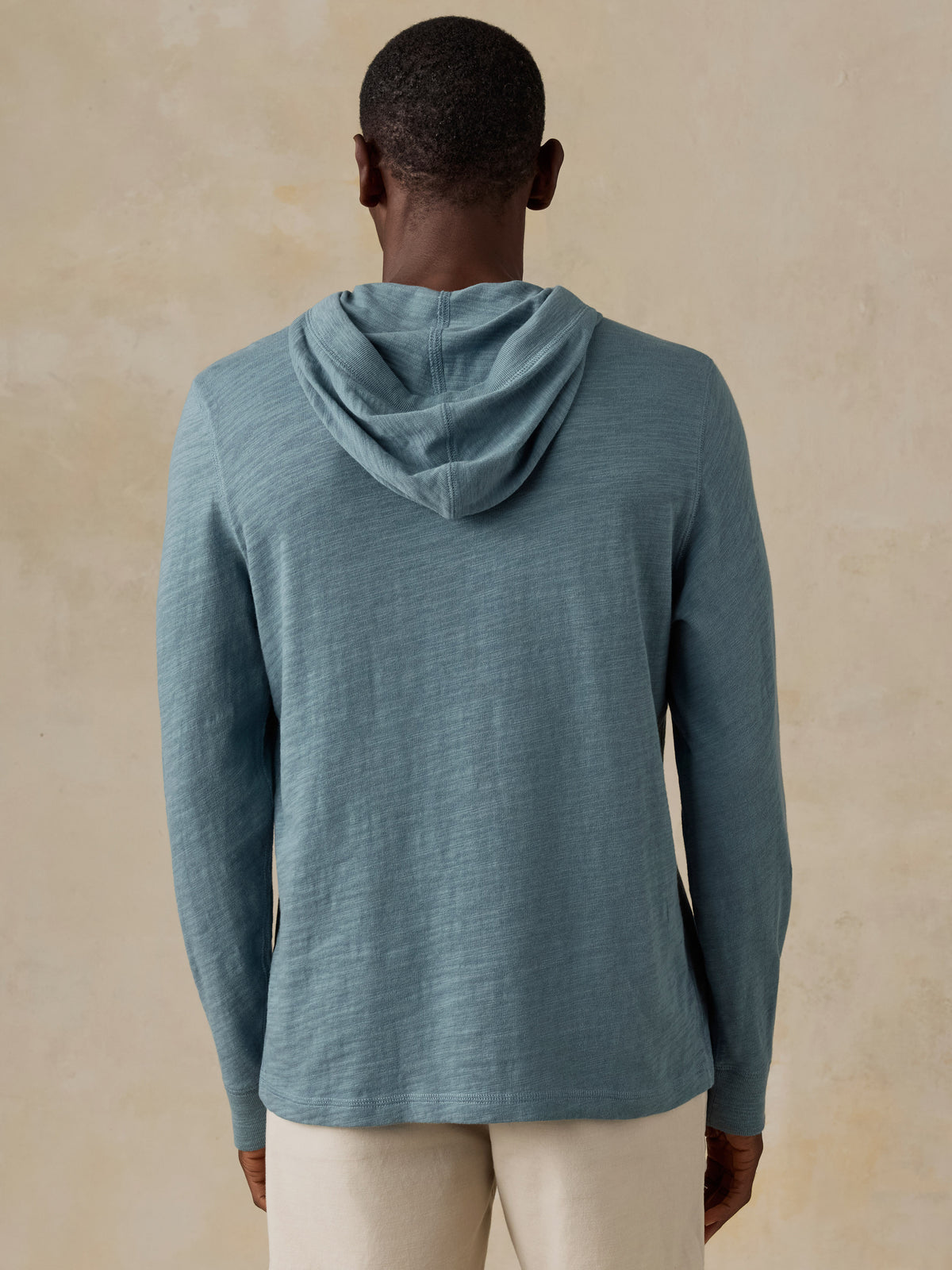 A man wearing a slate blue hooded long-sleeve shirt and cream pants., image 4 of 5