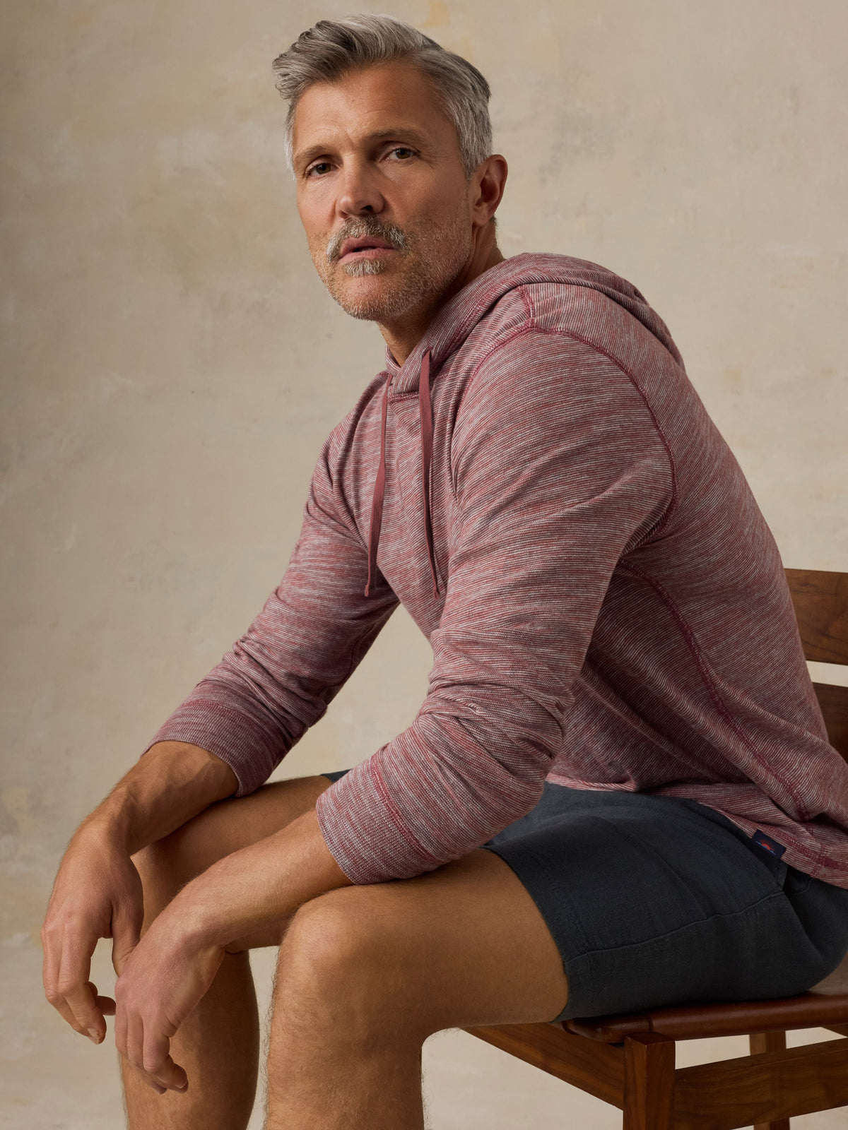Man sitting on a wooden chair wearing a red heathered hoodie and black shorts against a beige background, image 2 of 5