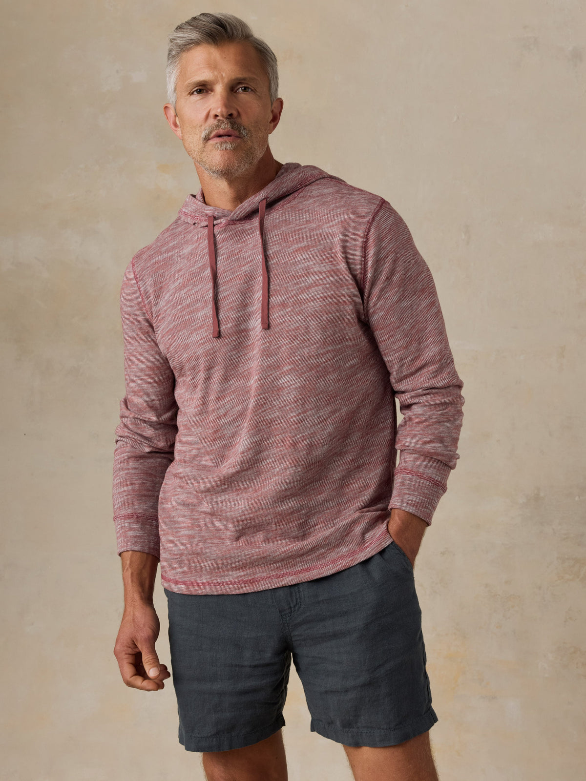 Man wearing a red heathered hoodie and black shorts against a beige background, image 1 of 5