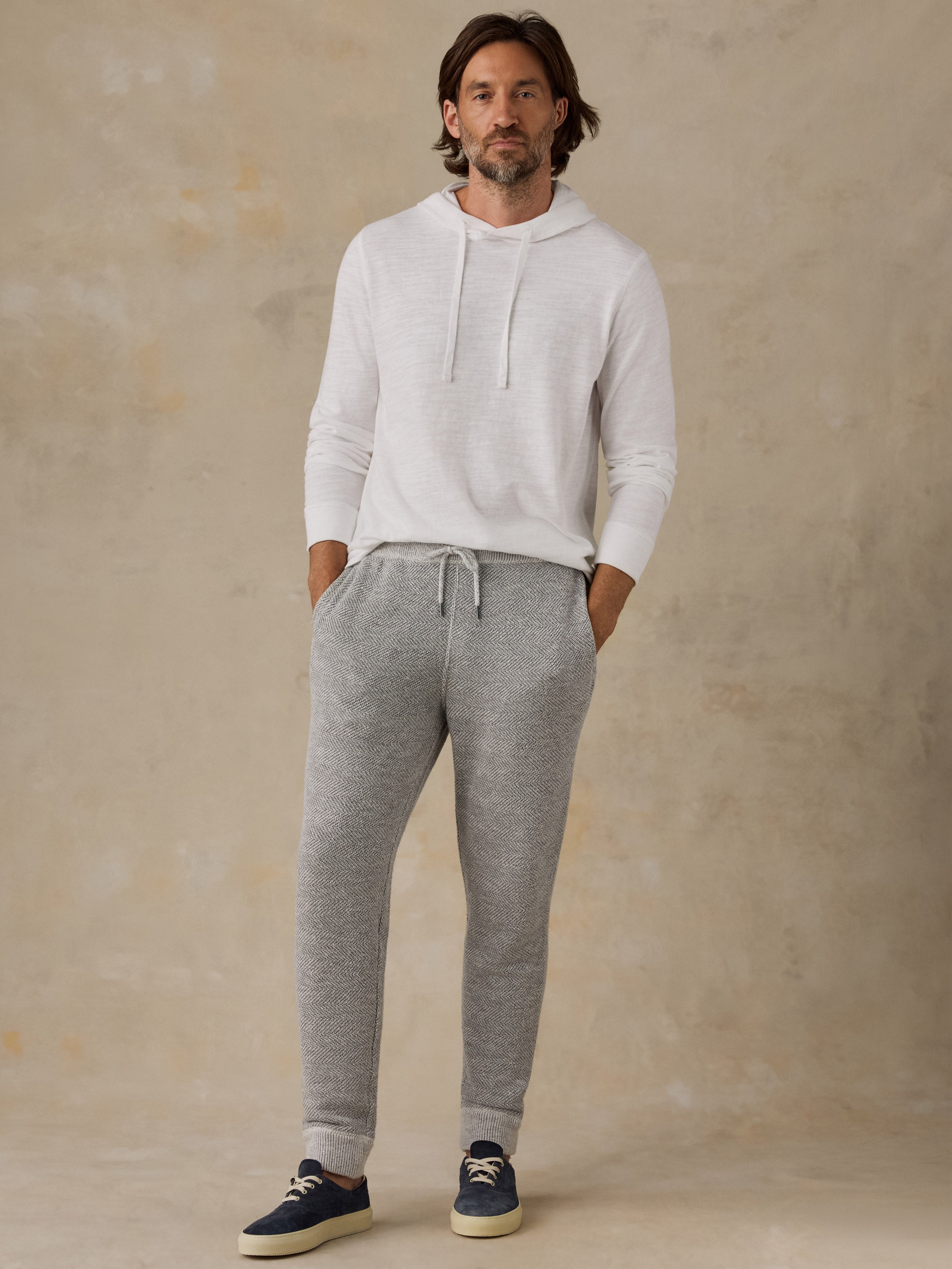 A man wearing a white long-sleeve hoodie and heather gray jogger sweatpants, standing against a textured beige studio backdrop.