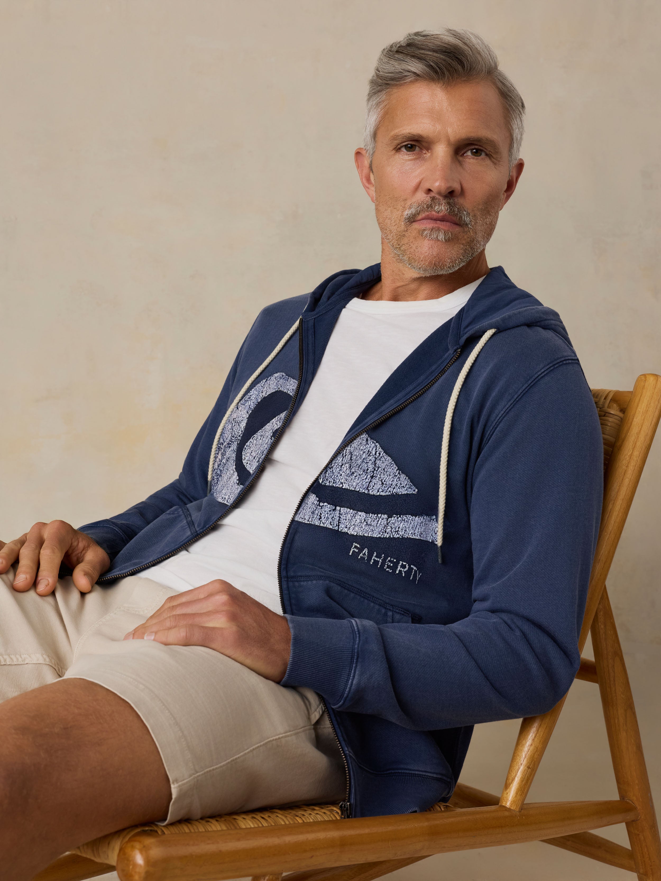 Man sitting on a wooden chair wearing a blue hoodie with a Faherty x Quiksilver and beige shorts against a beige background