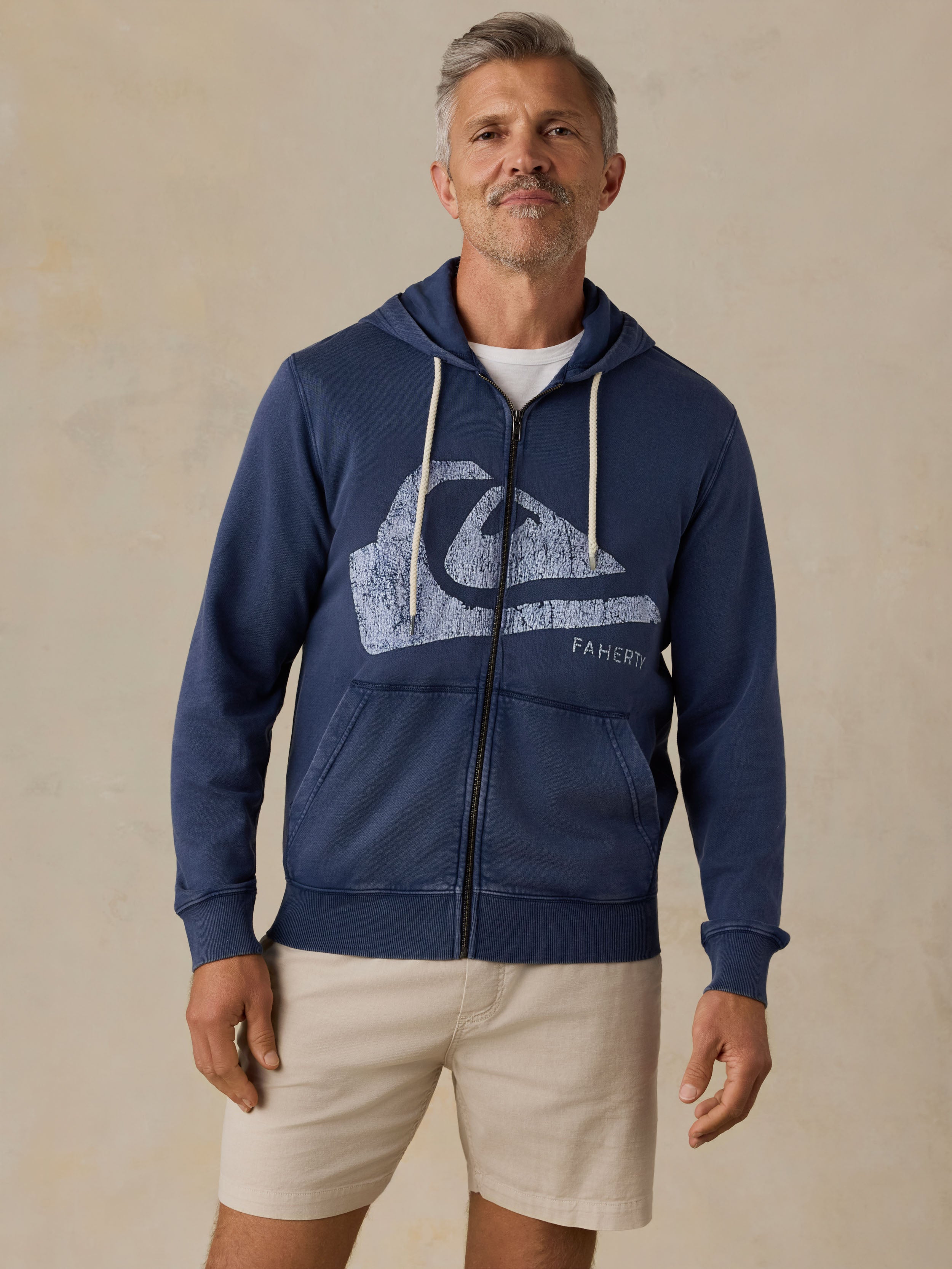 Man wearing a blue hoodie with a Faherty x Quiksilver and beige shorts against a beige background