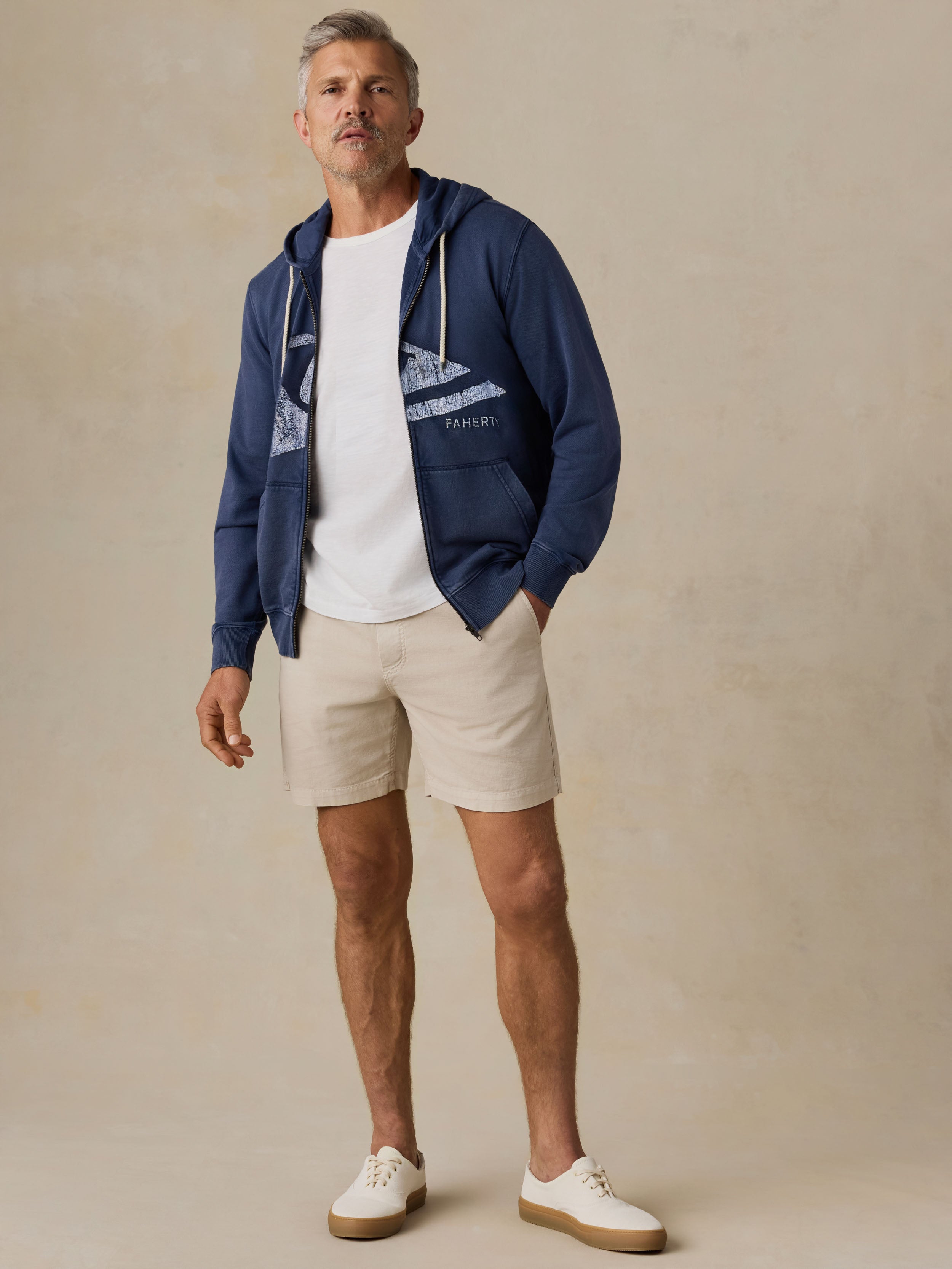 Man wearing a navy sweatshirt with Faherty x Quiksilver logo, white shirt, beige shorts, and white shoes on a beige background